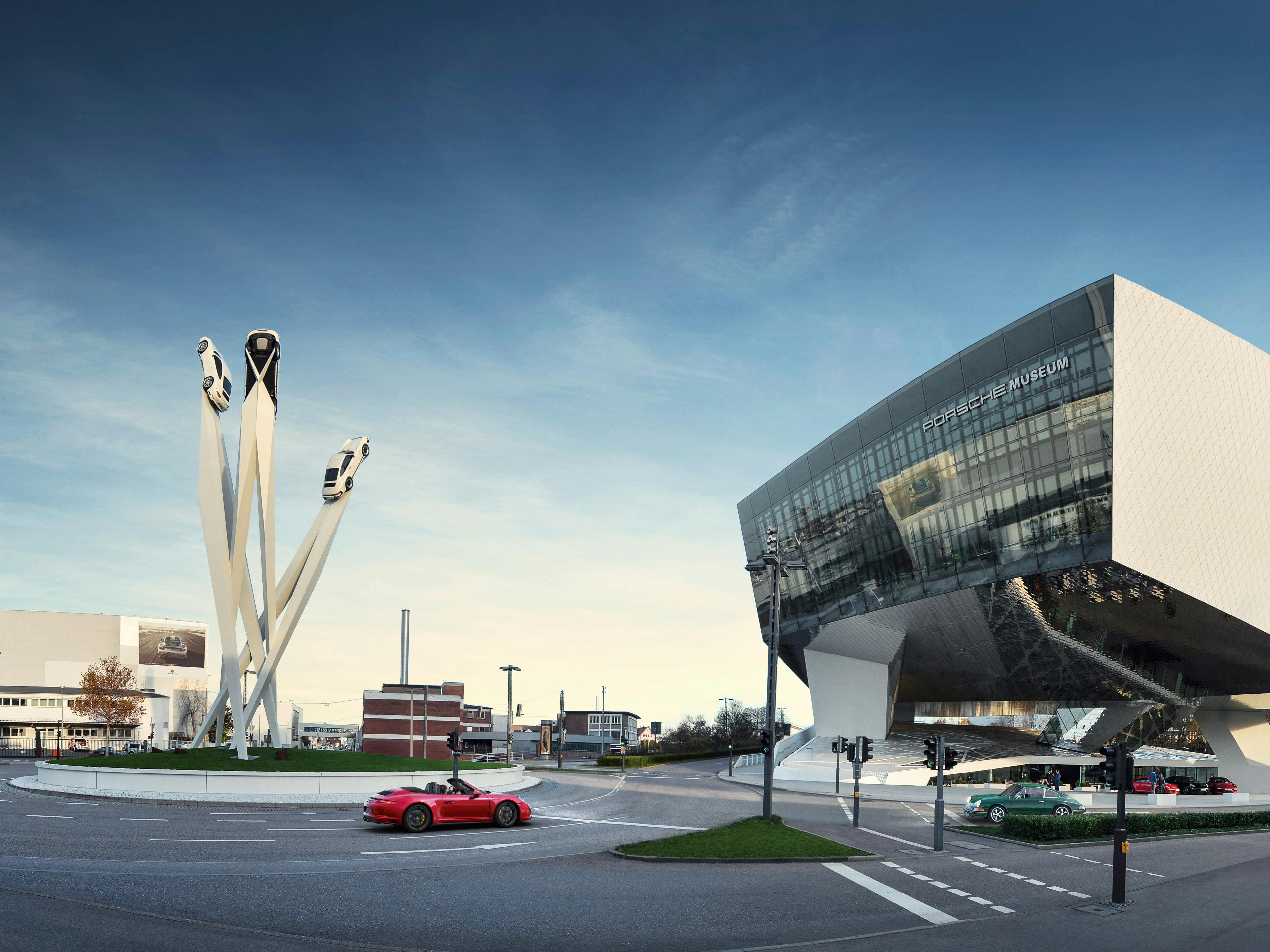 Das Porsche-Museum in Stuttgart mit einer markanten Skulptur im Vordergrund: Drei weiße Säulen, auf denen jeweils ein Porsche-Auto montiert ist, stehen auf einer runden, begrünten Verkehrsinsel vor dem modernen, kantigen Museumsgebäude.