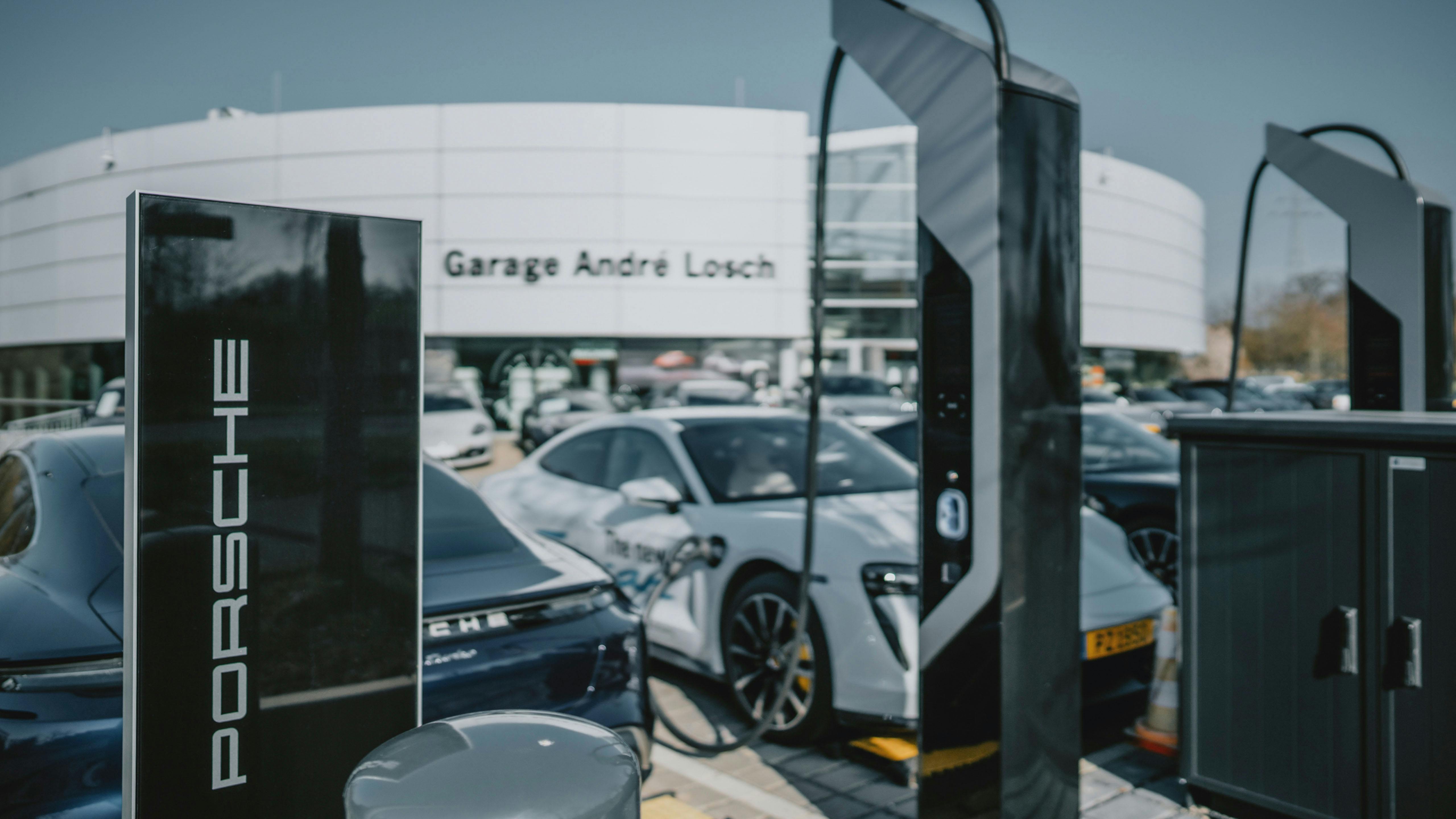 Rapproché d'une borne de recharge noire avec "Porsche" inscrit en lettres blanches, se tenant devant plusieurs voitures Porsche floues et dess bornes de recharge électriques, avec le Porsche Zenter Lëtzebuerg visibile en arrière-plan.