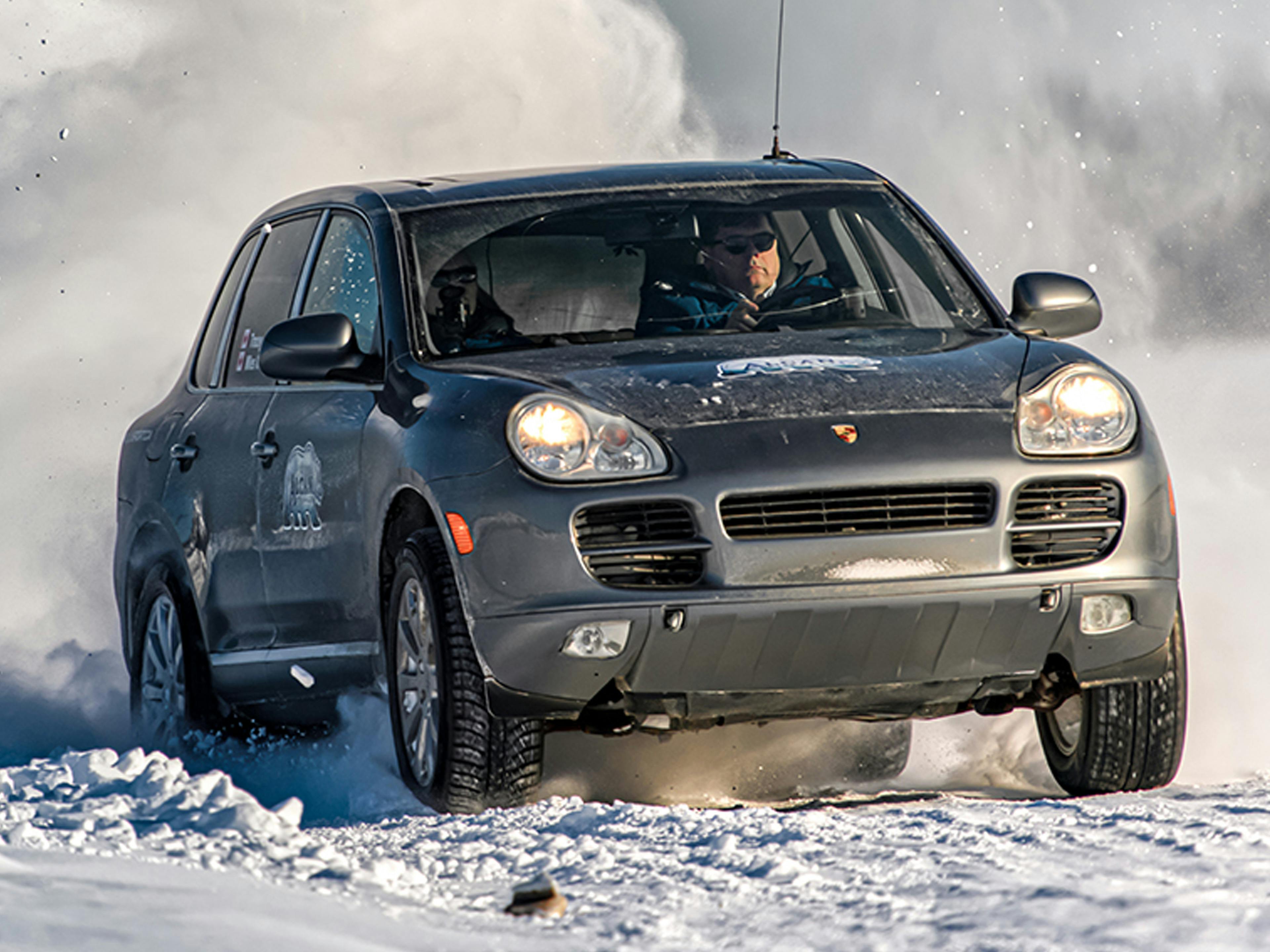 Calgary – A grey Porsche Cayenne powers through a snowy landscape, kicking up a cloud of snow behind it.