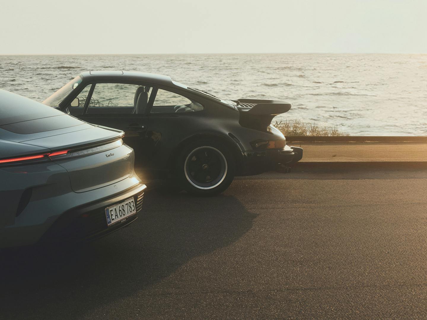 A modern Porsche and a classic 911 Turbo with its distinctive whale tail spoiler, parked together on a coastal road at sunset with the sea behind them.