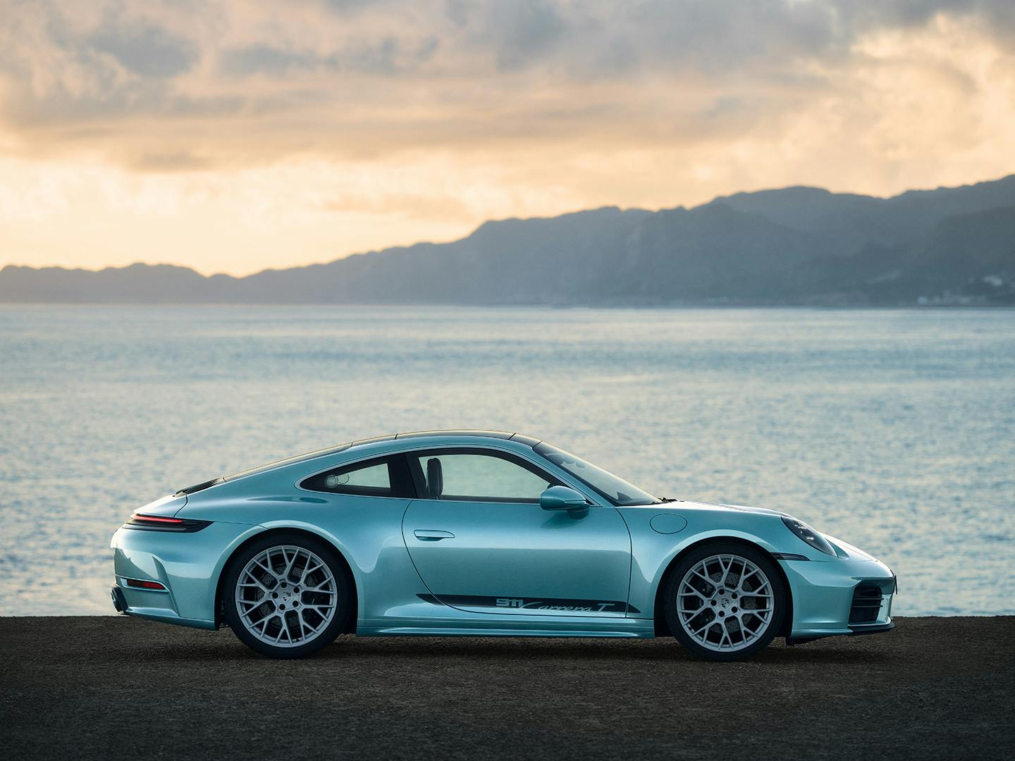 Detail of a turquoise Porsche 911 Carrera in side view, parked on a coastal road with mountains and sea in background.