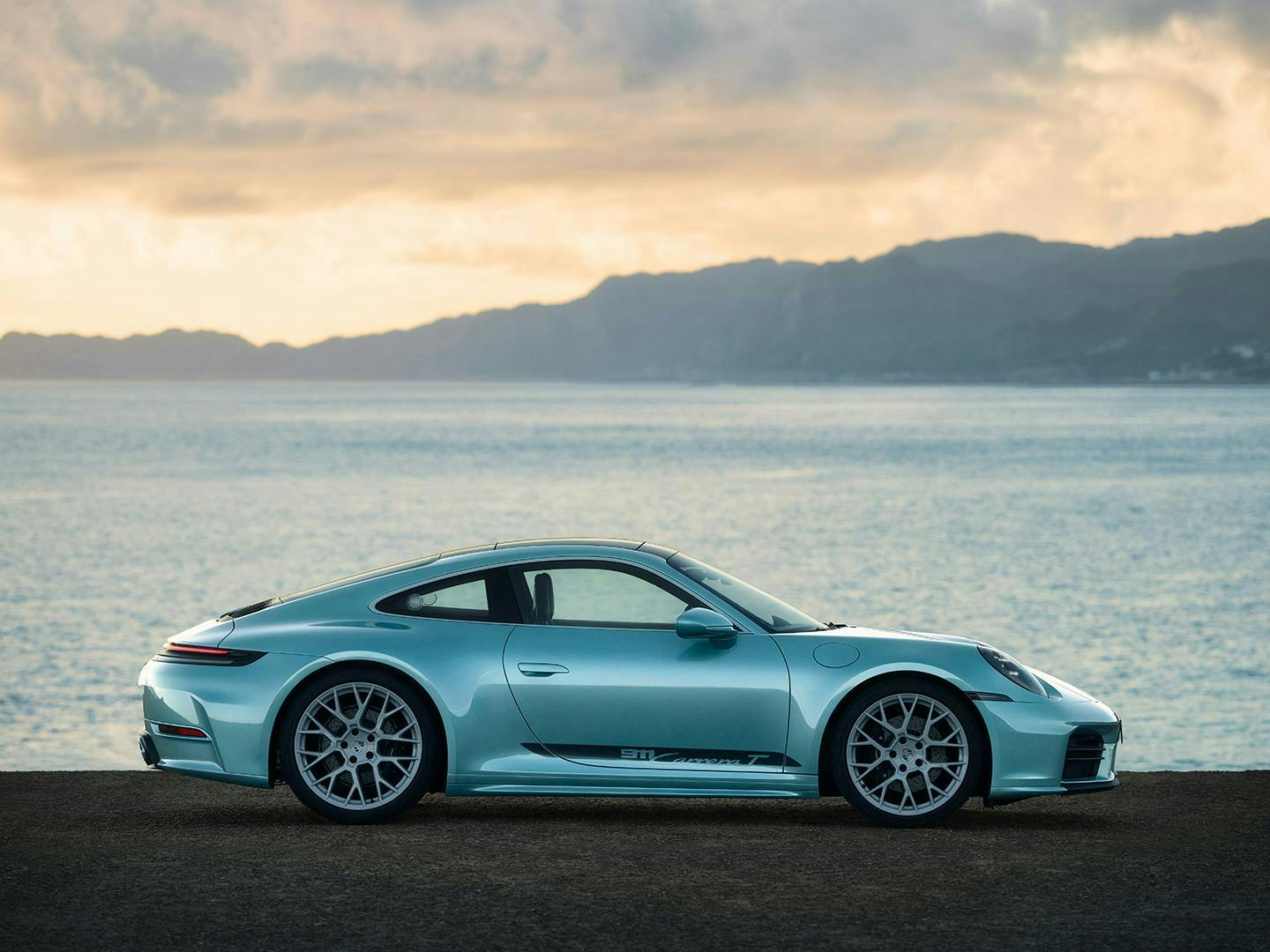 水綠色 Porsche 911 Carrera 側視細節照，停在海岸公路上，背景為山脈與大海。