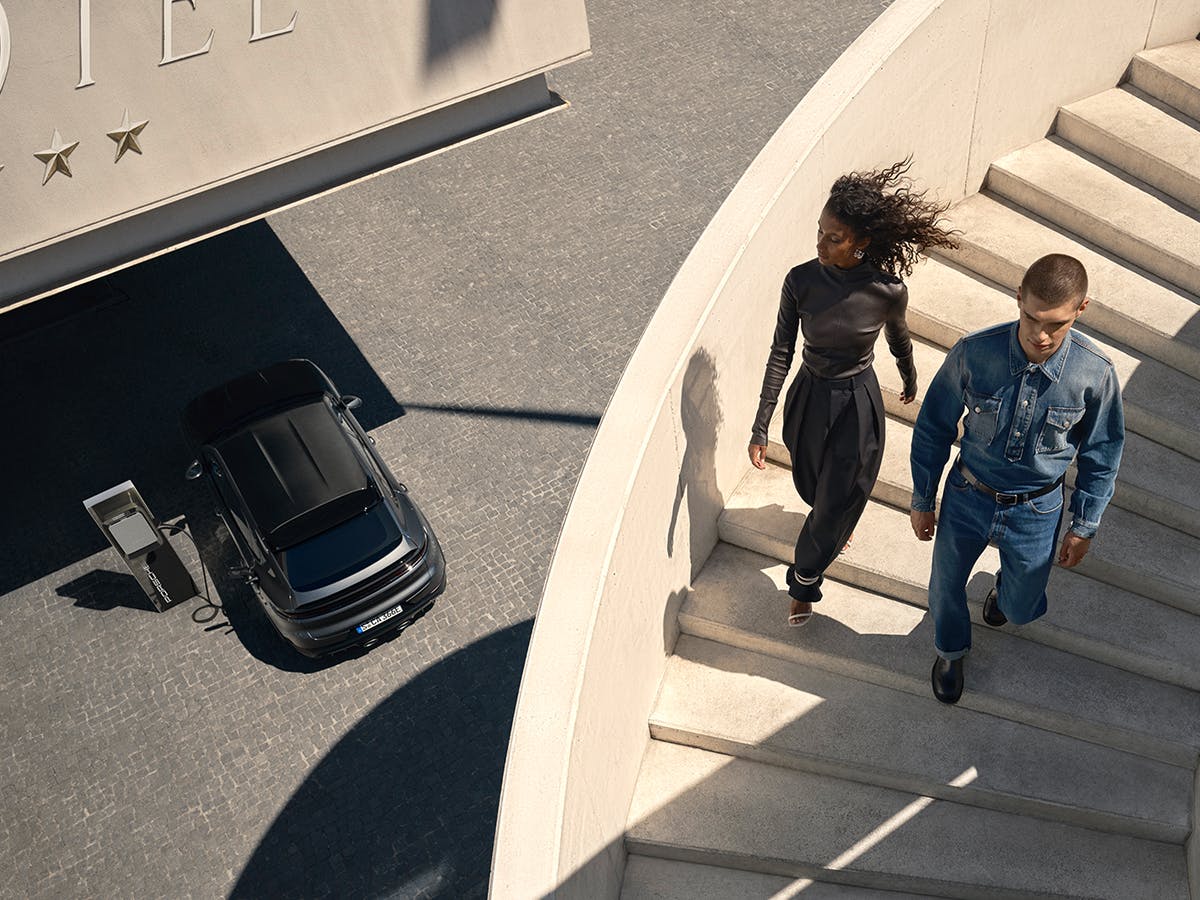 A woman and a man walking down a staircase. A Porsche Macan is charging at a charging pedestal below.