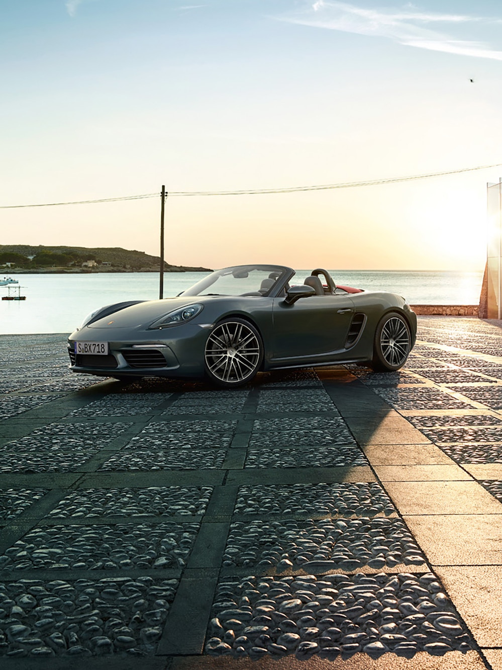 A grey metallic Porsche 718 Boxster vehicle parked outside with a sea view in the background.