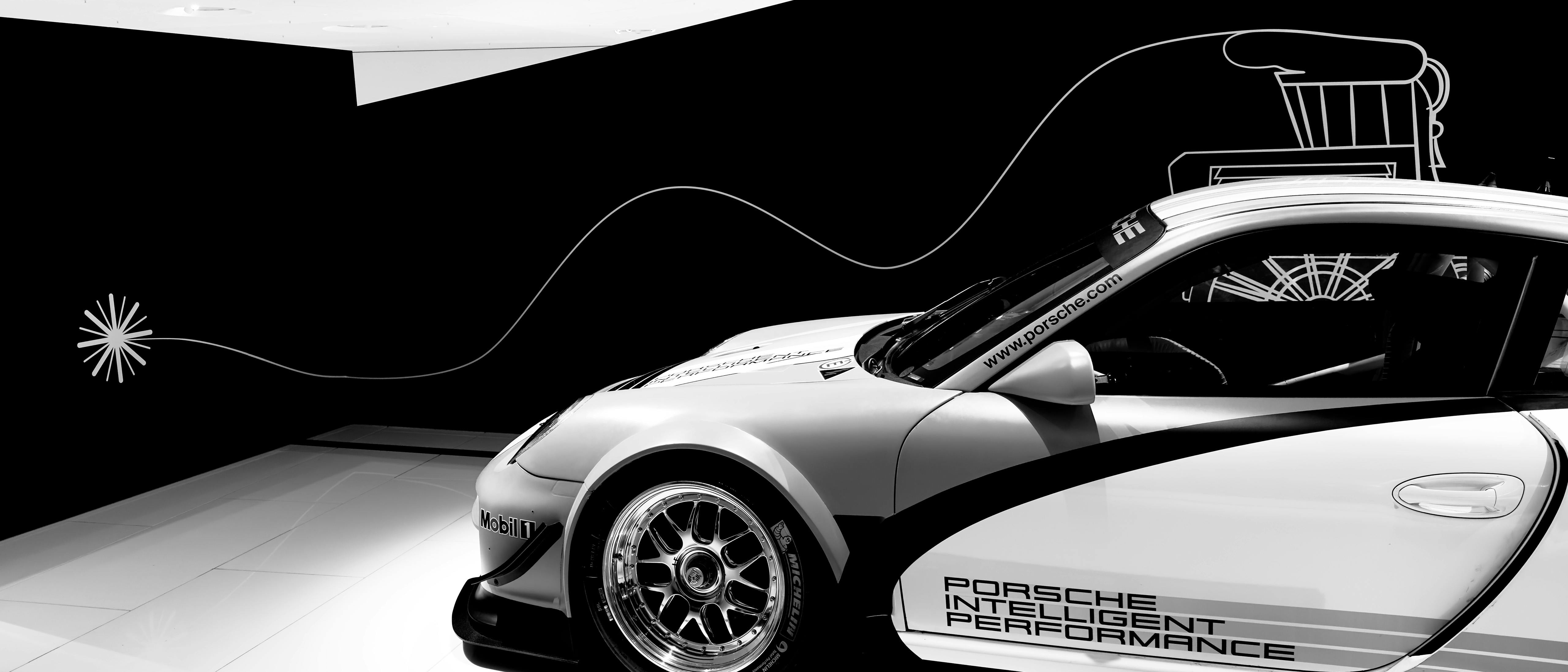 Teilansicht eines Porsche‑Rennwagens vor schwarzer Ausstellungswand mit grafischen Linien und technischen Illustrationen im Hintergrund.