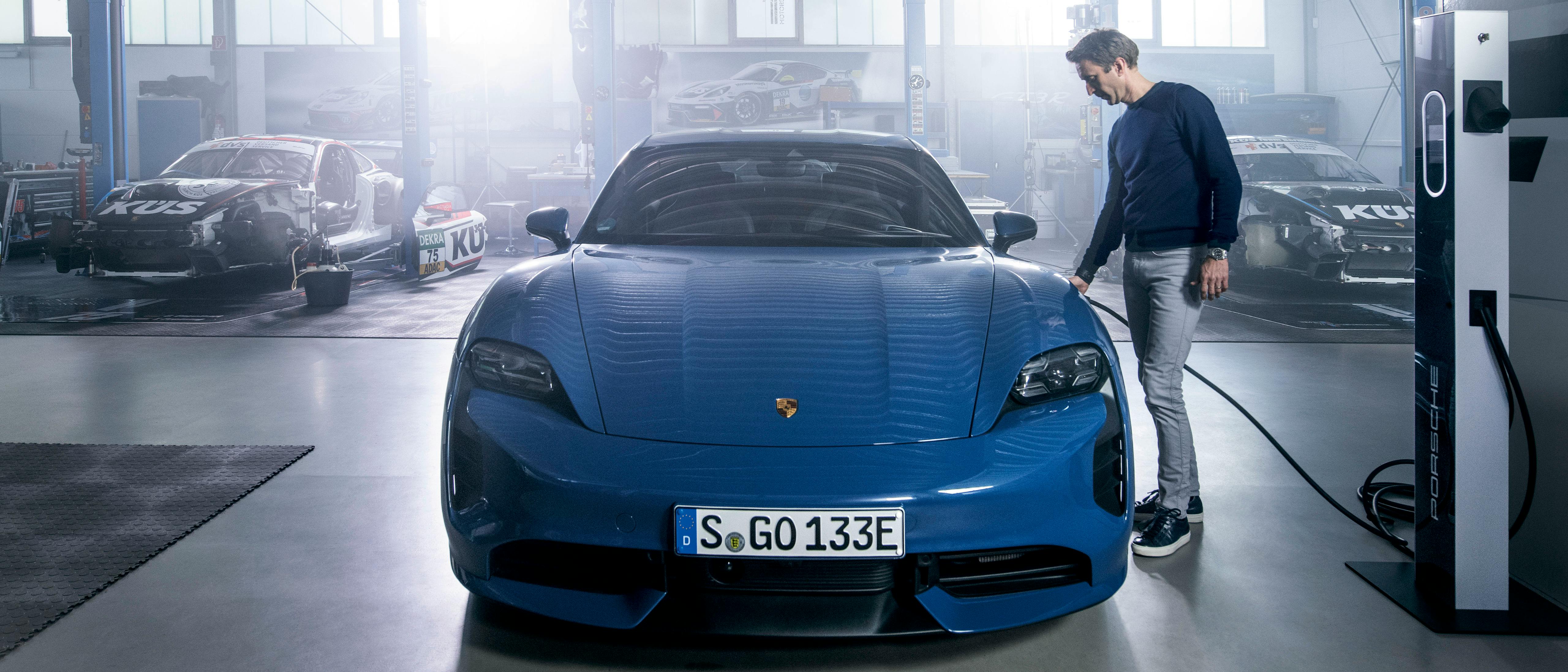 Frontal view of a Porsche Taycan. A man is standing on the right side at the charge port and is charging the vehicle.