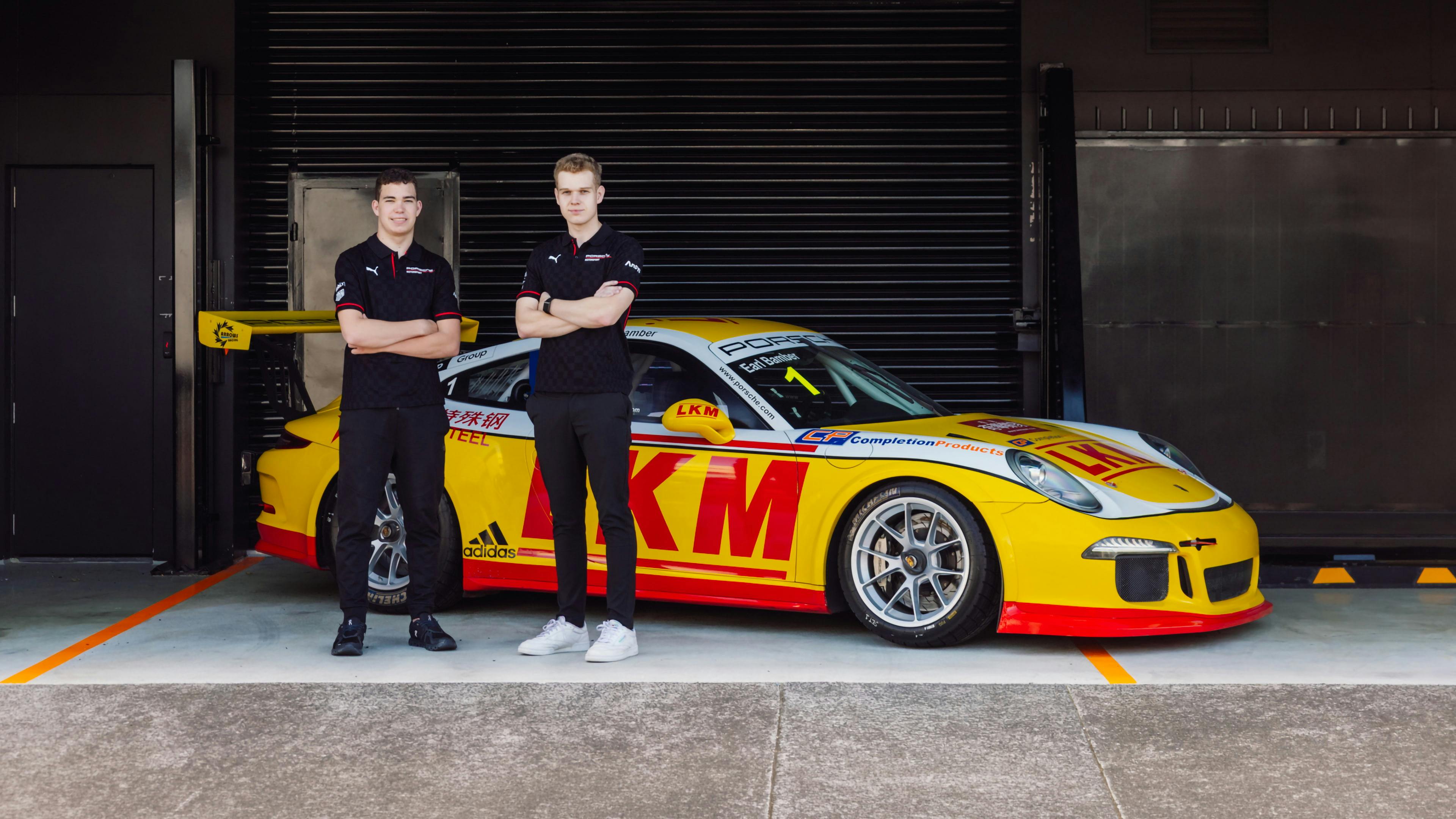 Two people standing in front of a yellow Porsche race car inside a garage, with arms folded and a roller door behind them.