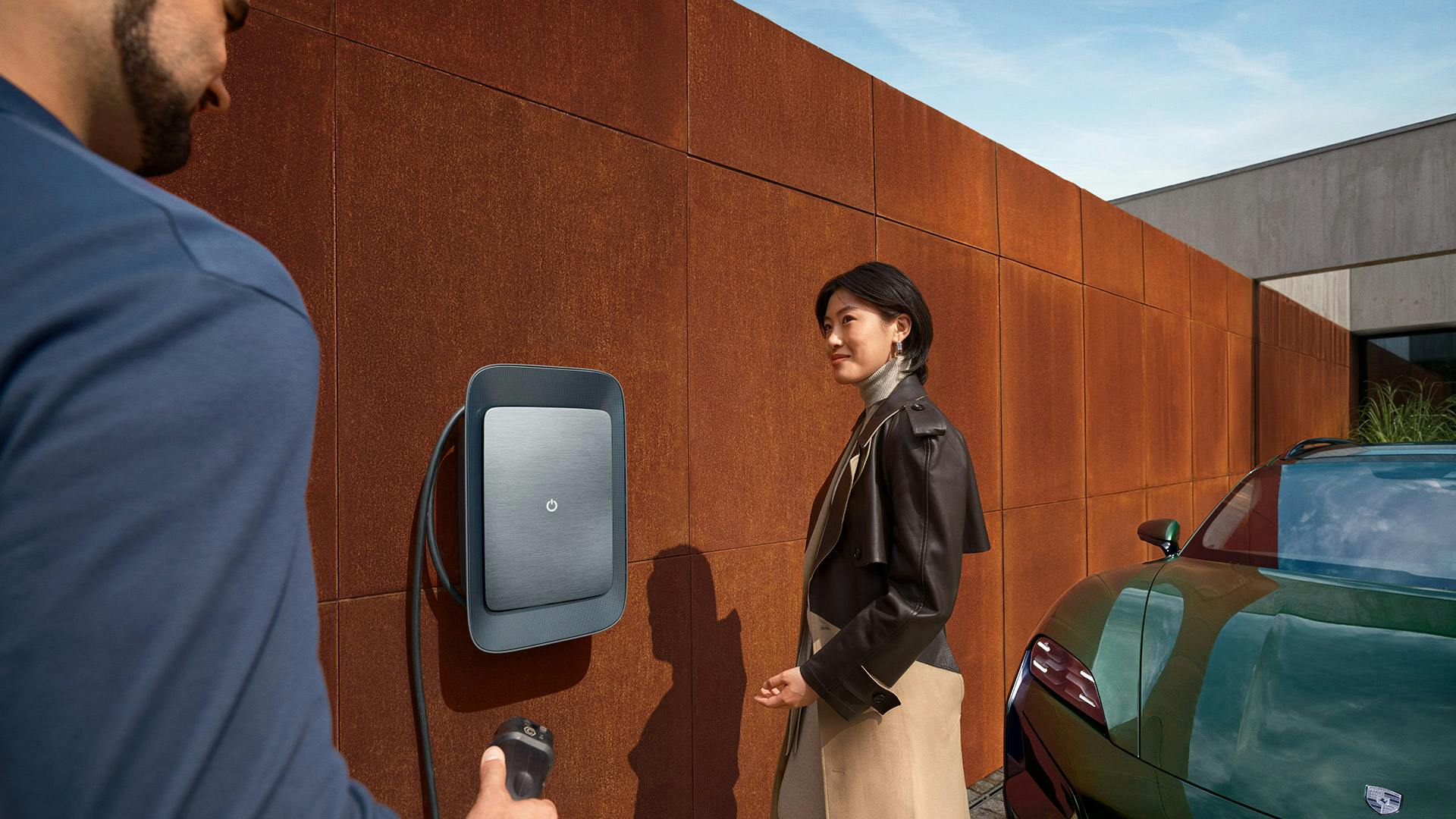 A man and woman stand beside a green Porsche Taycan charging at home with a Porsche Wall Charger against a modern facade.