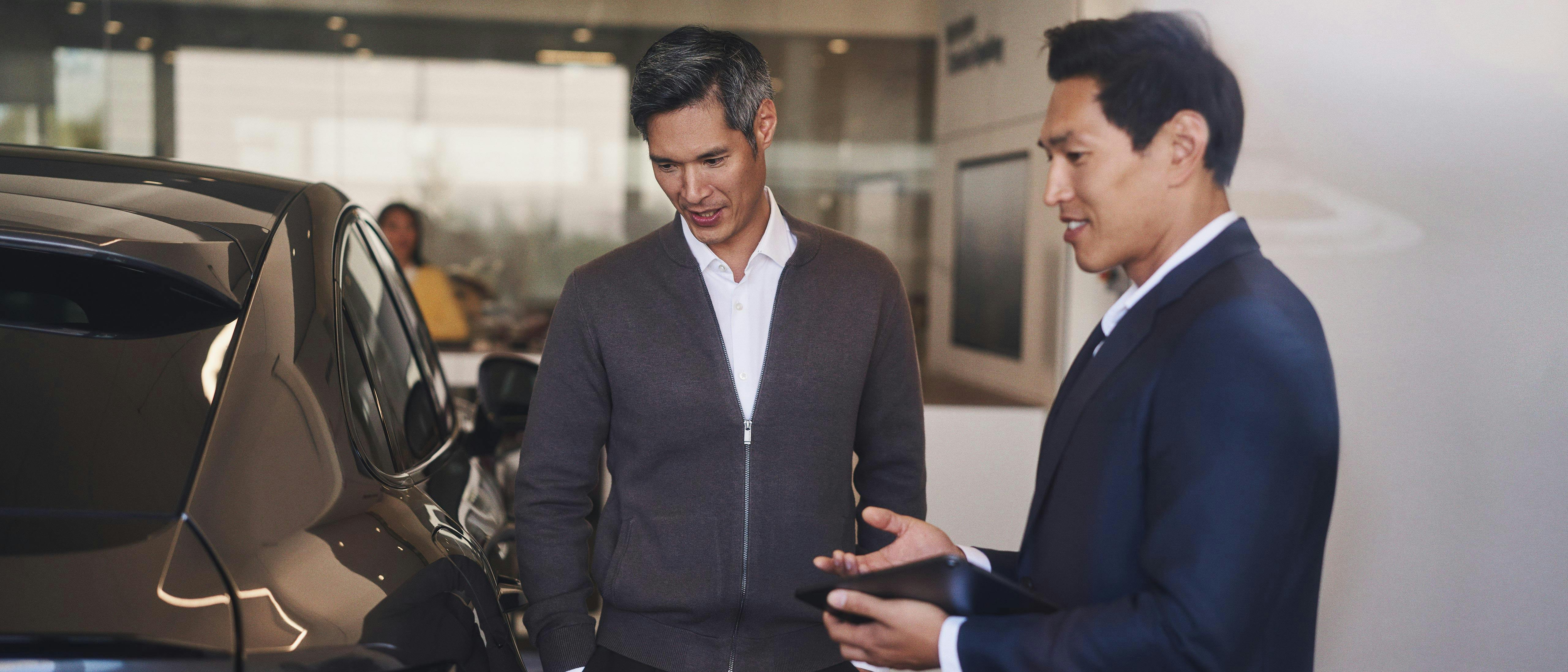 Two men in a car dealership. Porsche employee in a blue suit holds an iPad in his hand and presents a Porsche car to the customer.