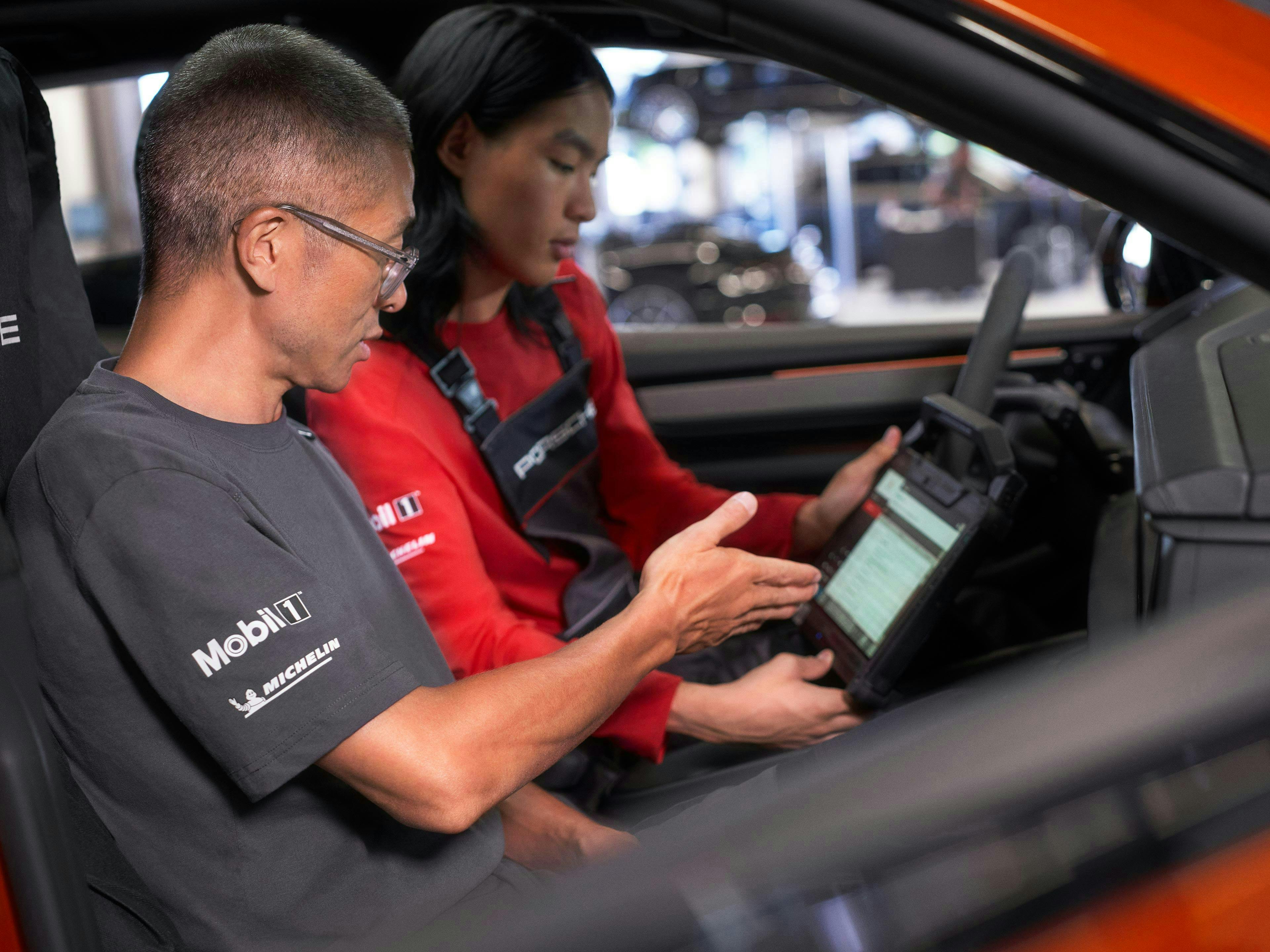 Two Porsche mechanics are sitting in a papaya-colored Macan and looking at the Porsche tester, with which they are currently conducting a vehicle diagnosis.