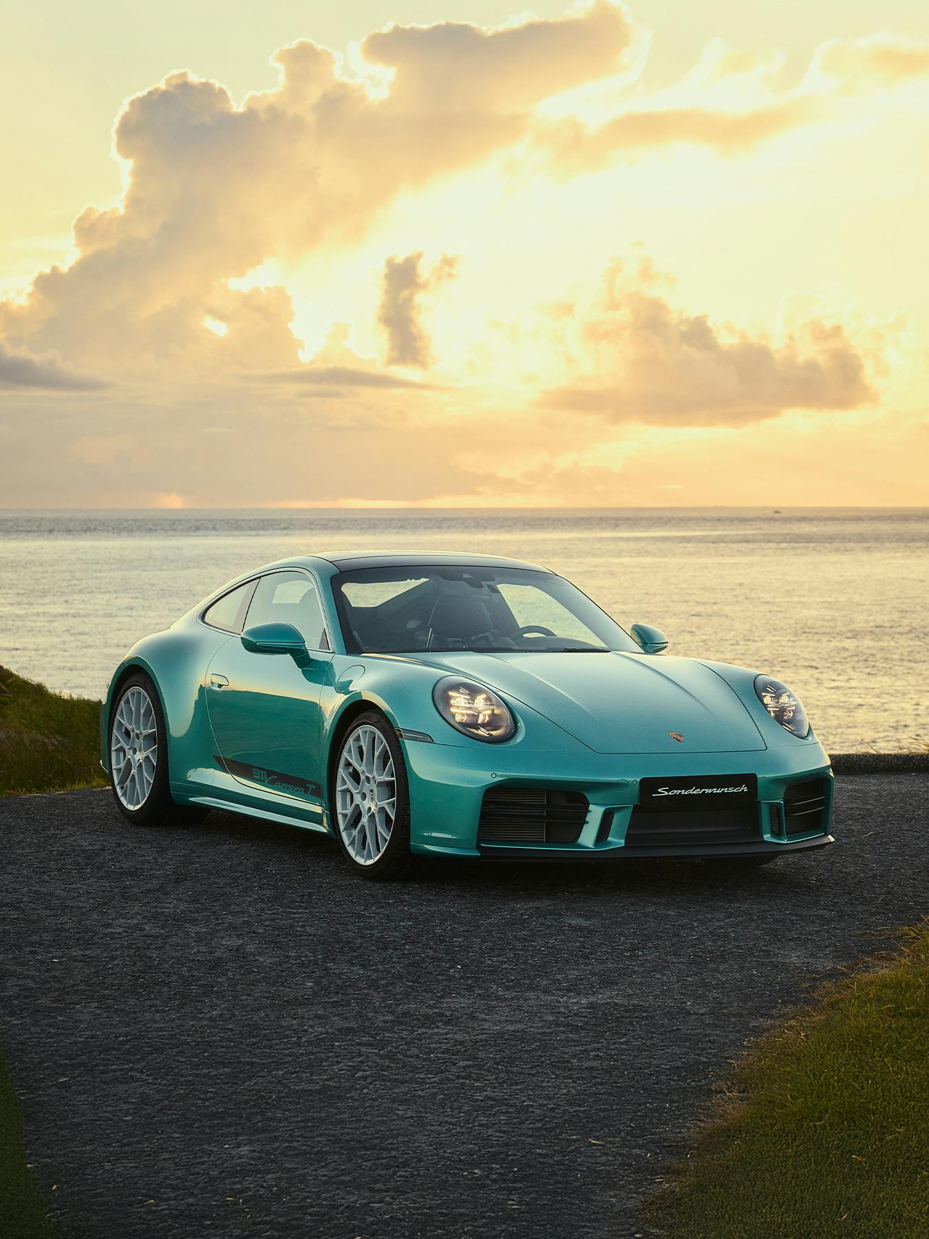 A turquoise Porsche 911 Carrera T parked on a coastal road at sunset, with ocean and glowing clouds behind.”