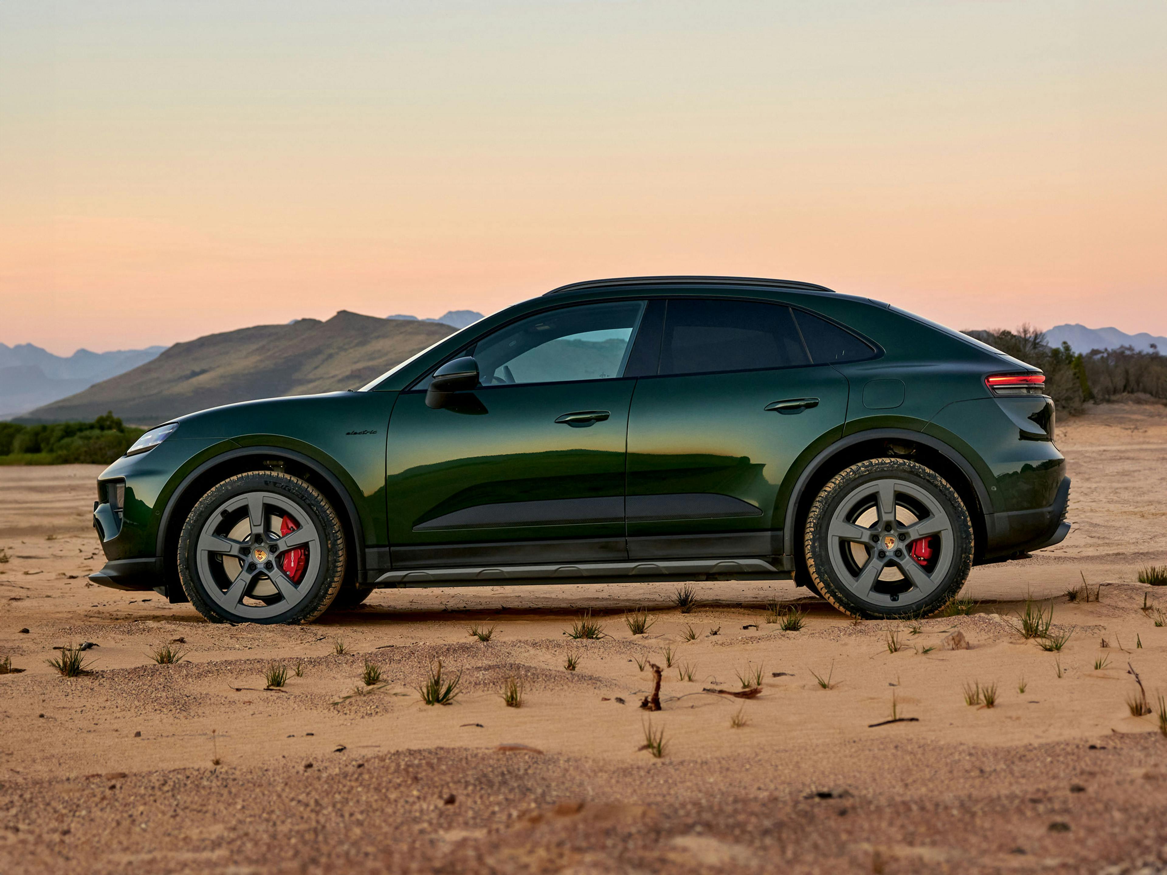 Side profile of green Porsche Macan 4S Electric parked on sand with mountainous backdrop.