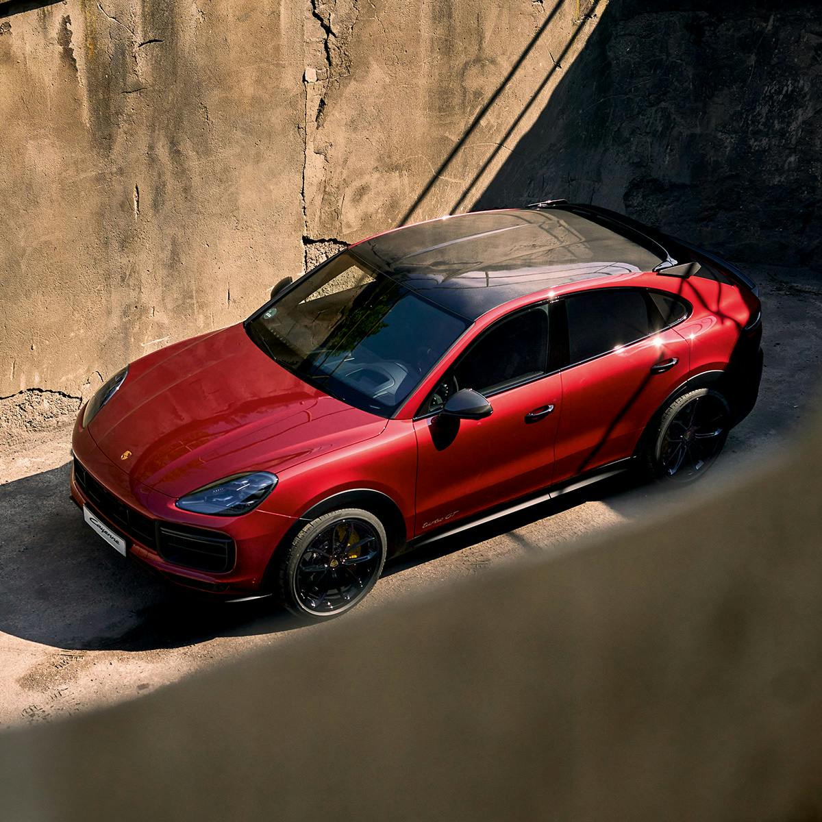A red Porsche Cayenne Coupe is parked in a sunlit area, surrounded by tall concrete walls. The car features a sleek, sporty design with black accents, tinted windows, and a panoramic glass roof.