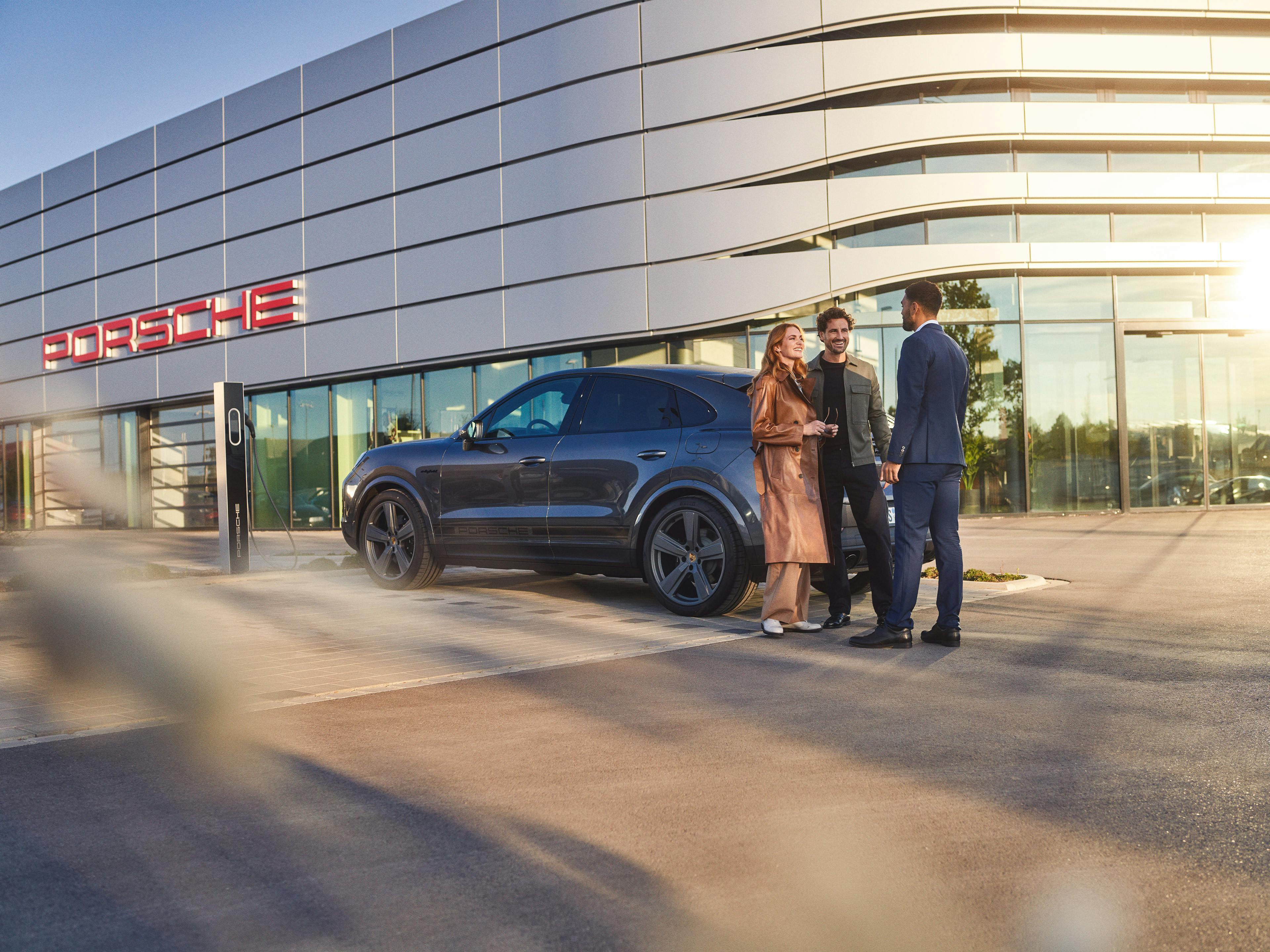 Un couple et un conseiller du Centre Porsche restent ensemble près d'un véhicule Porsche devant l'entrée du Centre Porsche.