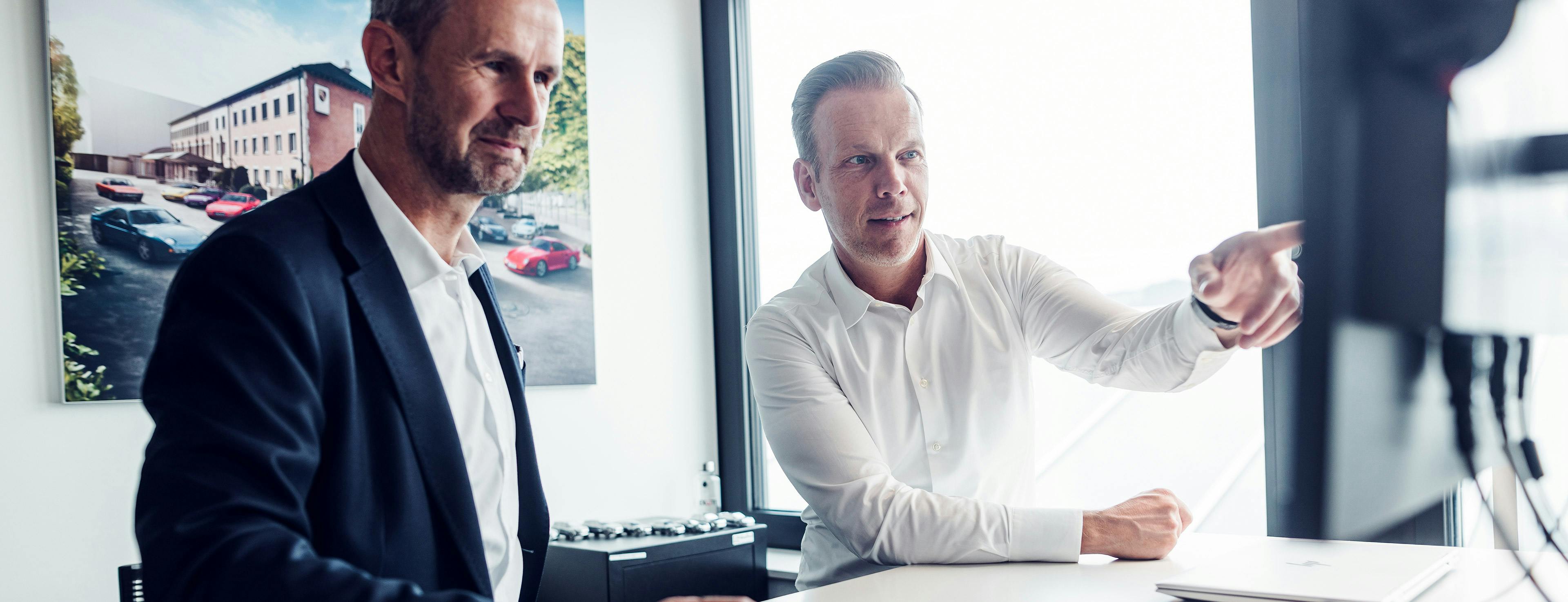 Zwei Porsche Mitarbeiter sitzen an einem Schreibtisch und schauen auf einen Monitor, einer von beiden zeigt auf etwas.