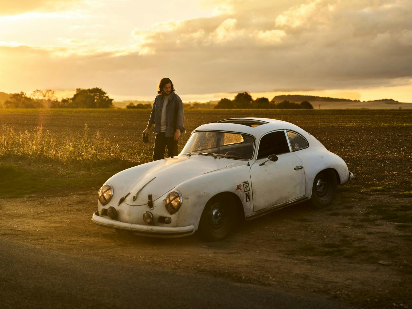 A man is standing next to a white Porsche classic car in the countryside during sundown.