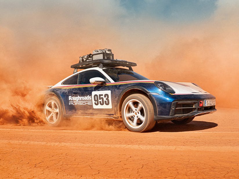A Porsche 911 Dakar drives through a dusty desert landscape; a roof cage with securely lashed equipment is mounted on the roof.