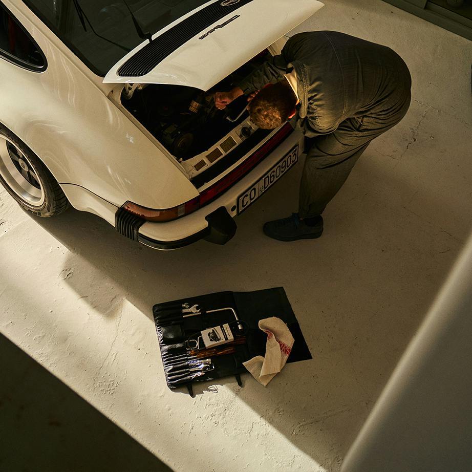 Een monteur sleutelt in de garage aan de motor van een Porsche 911 Classic model in wit met de motorkap open.