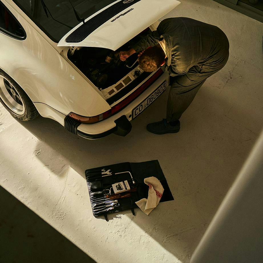 Un mécanicien effectue des opérations de vissage sur le moteur d’un modèle Porsche 911 (Porsche Classic) en Blanc avec le capot moteur ouvert dans le garage.