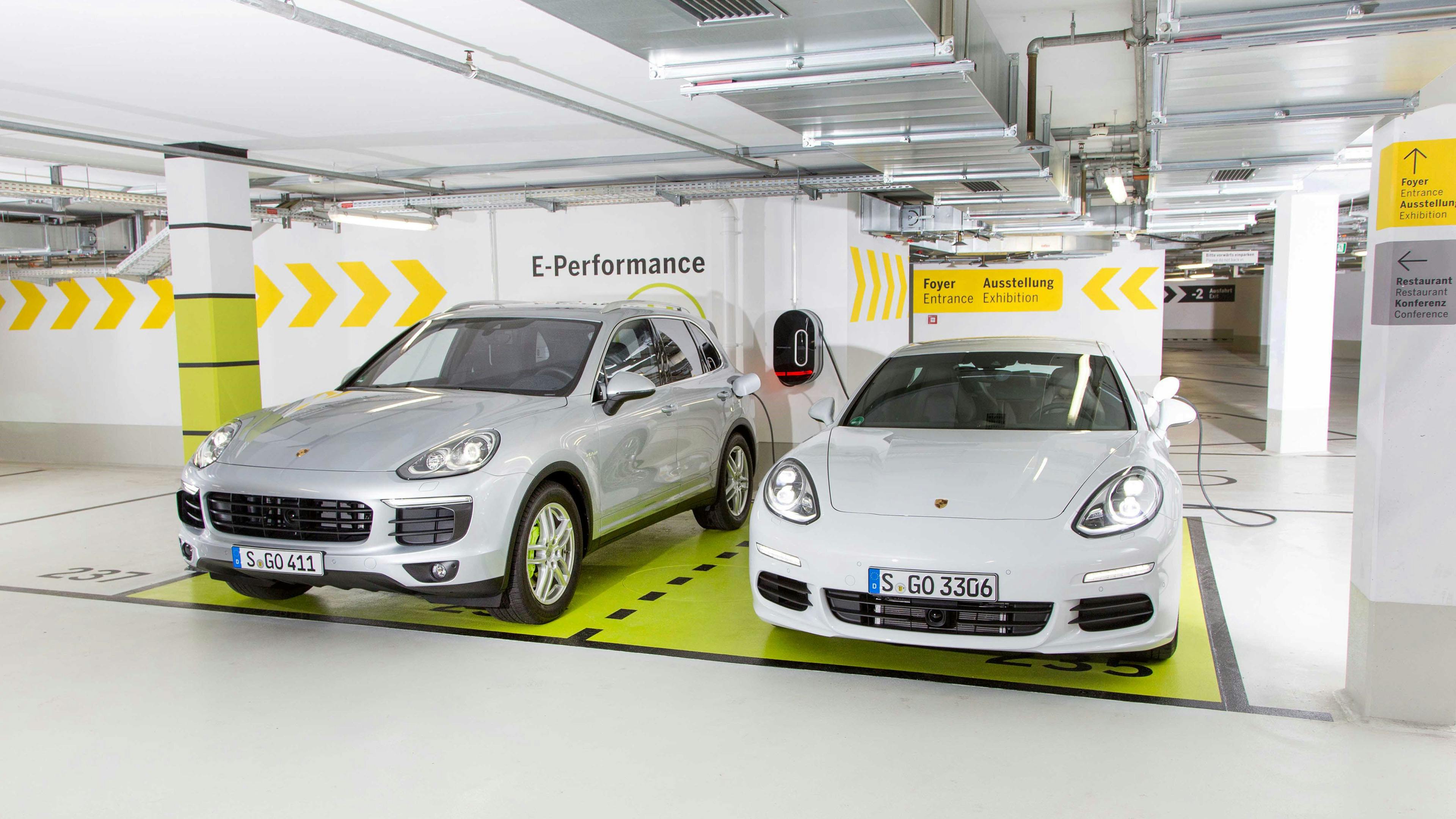 Zwei Porsche E‑Hybrid Fahrzeuge stehen nebeneinander auf gekennzeichneten E‑Performance‑Parkplätzen in einer hellen Tiefgarage. Beide Fahrzeuge sind an Ladestationen angeschlossen.