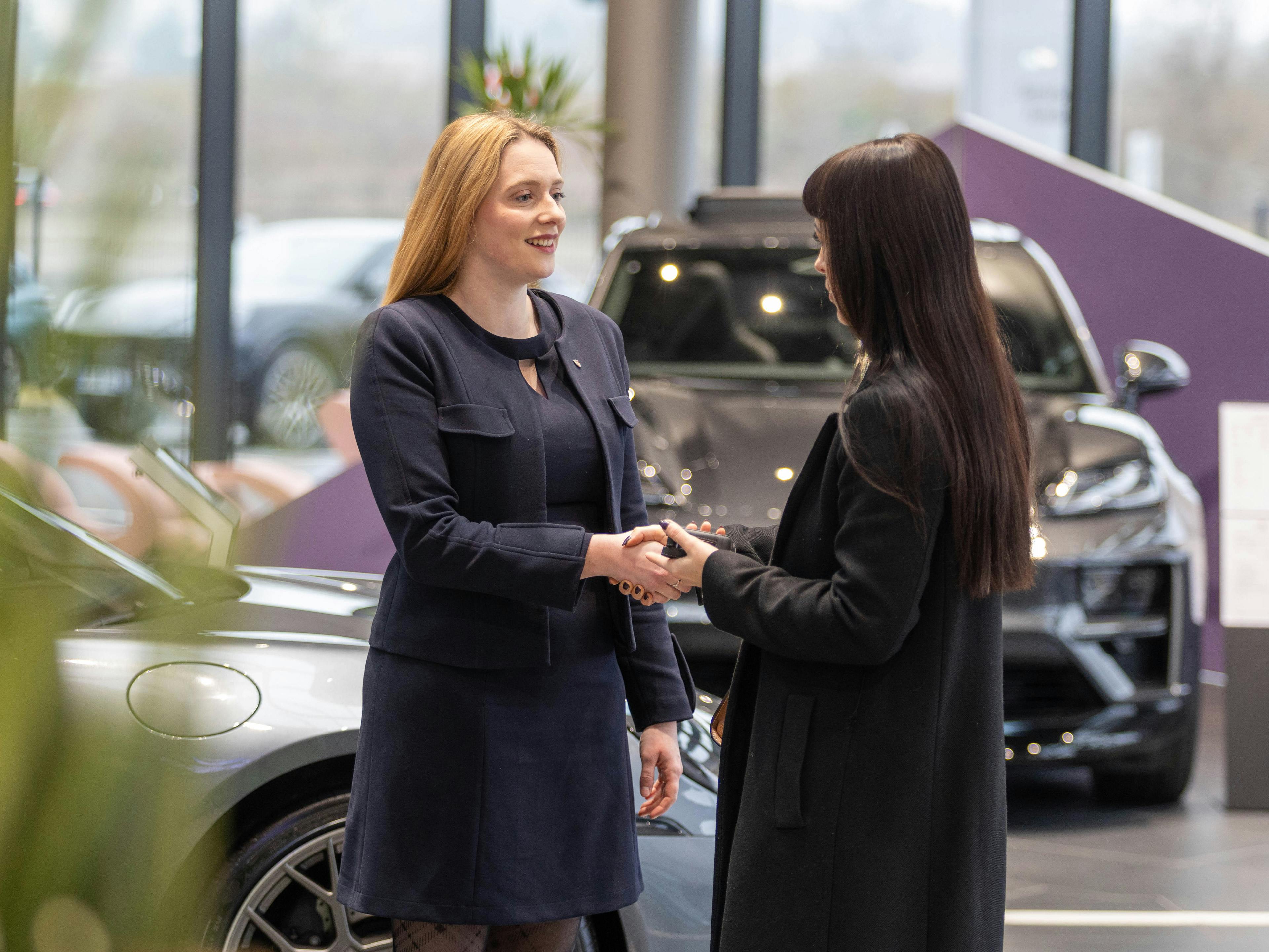 Une collaboratrice de Porsche salue une cliente en lui serrant la main. Toutes deux se tiennent devant un véhicule Porsche gris dans la salle d'exposition d'un Centre Porsche.