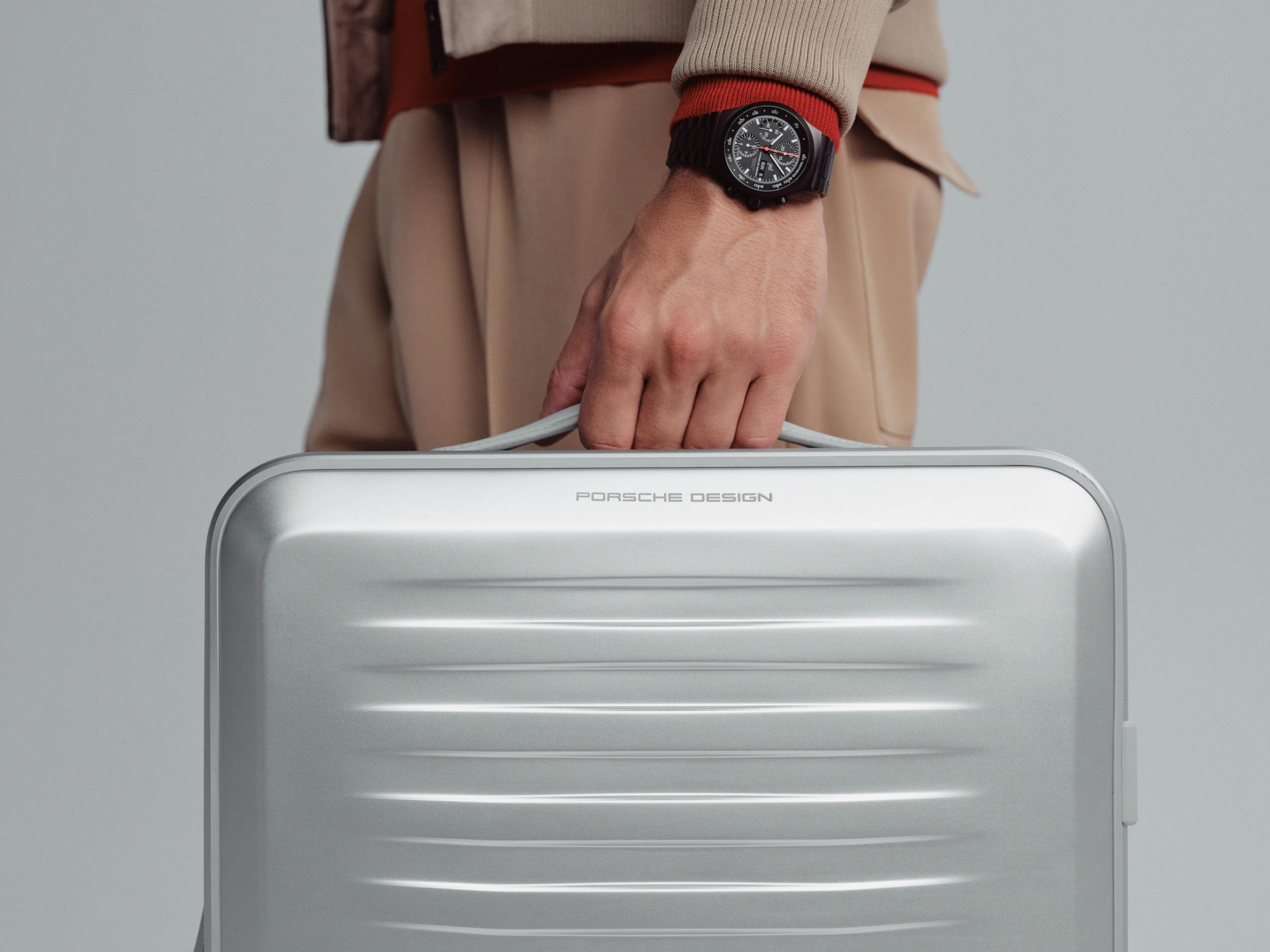 Ein Mann in beigefarbener Kleidung hält einen silbernen Koffer mit der Aufschrift „Porsche Design“ in der Hand. Am Handgelenk trägt er eine schwarze Armbanduhr mit komplexem Zifferblatt. Der Hintergrund ist neutral grau und minimalistisch gehalten.