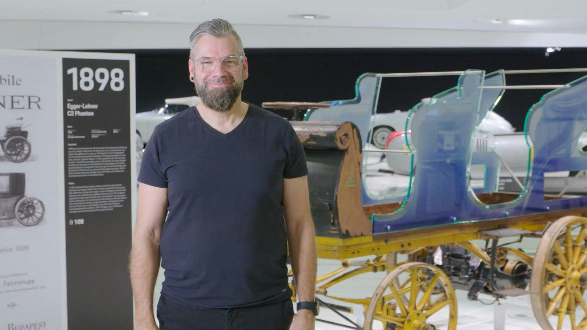 Ein lächelnder Mann in dunklen Klamotten, der im Porsche Museum vor einem Porsche Modell von 1898 steht.