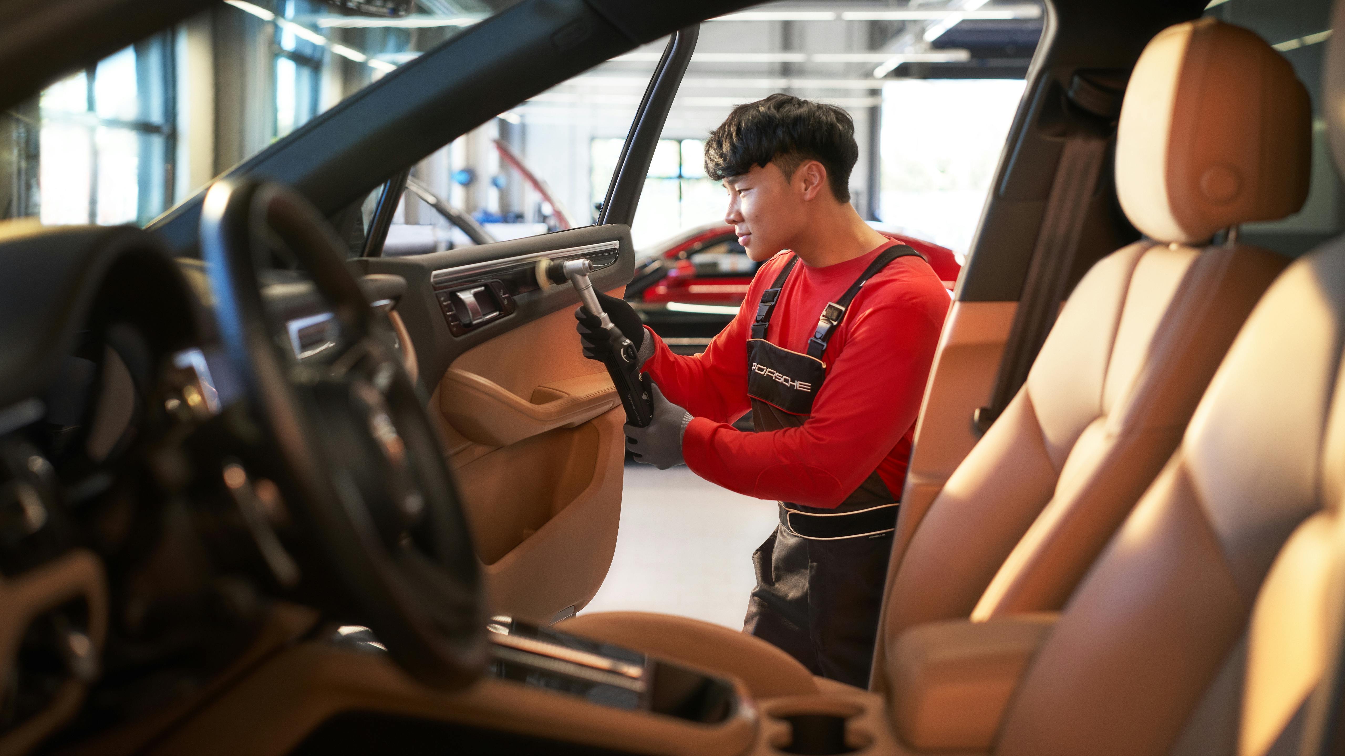 Porsche employee carefully cleaning the interior of a car with beige leather seats in the workshop.