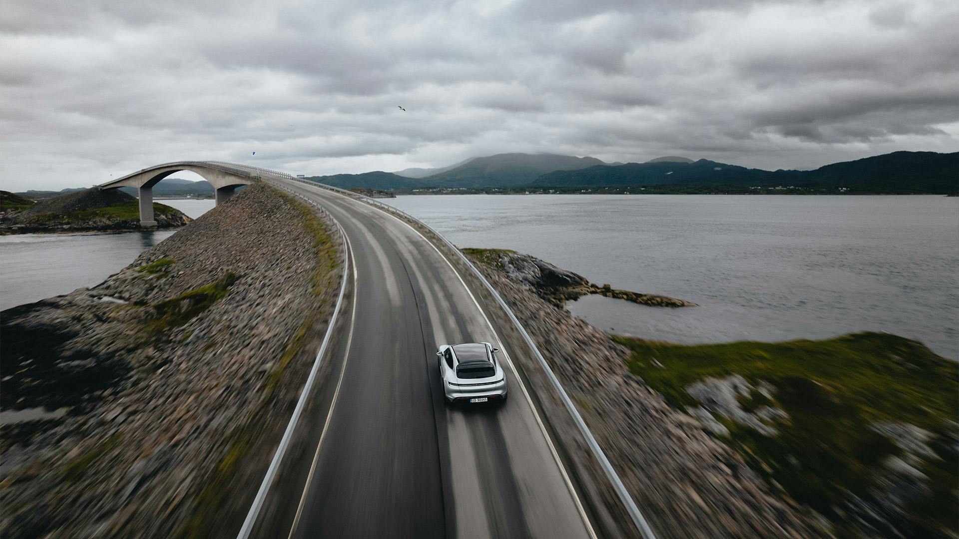 Un vehículo Porsche se acerca a un puente. Al fondo: las montañas y la inmensidad del agua.