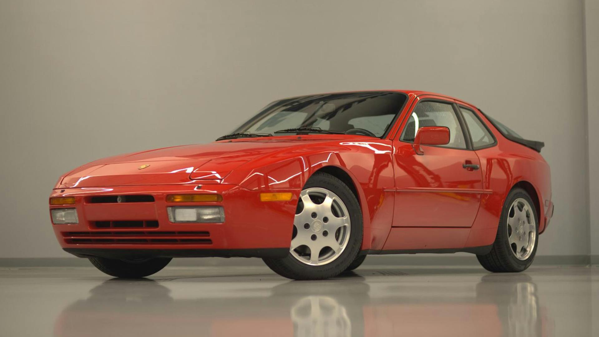 A red sports car with a sleek design and pop-up headlights is parked indoors against a gray wall.