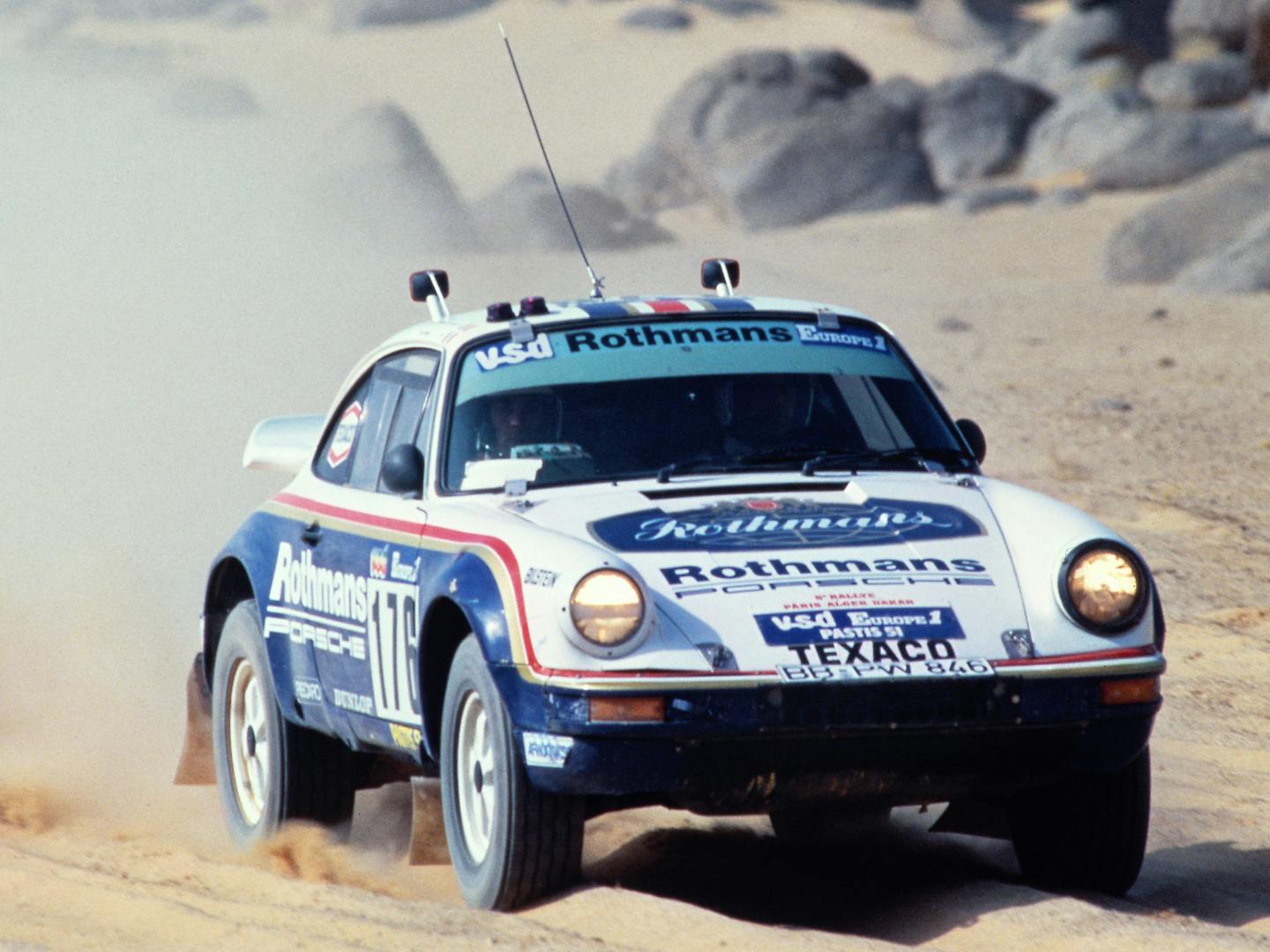 A white and blue Porsche 911 SC 4x4 rally car kicking up dust as it races across the desert.