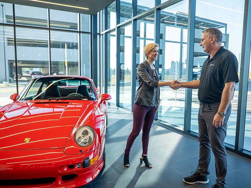 Factory Restoration employee standing next to a red classic Porsche, shaking hands with a customer.