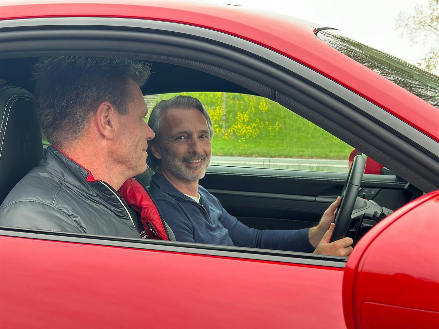 Close-up van het interieur van een rode Porsche 911 tijdens een proefrit. Een lachende man achter het stuur en een instructeur in een Porsche-bodywarmer genieten. Zij genieten van de rijervaring. De focus ligt op de luxe afwerking van het dashboard en de sportstoelen.