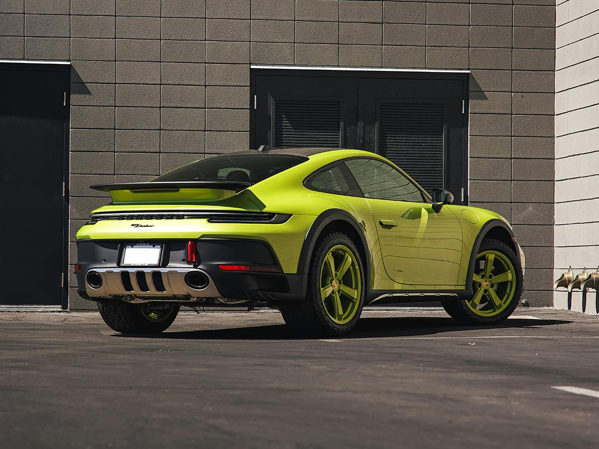A 911 Dakar coloured in Peridotmetallic with wheel rims in the same colour from behind in front of a brick building.