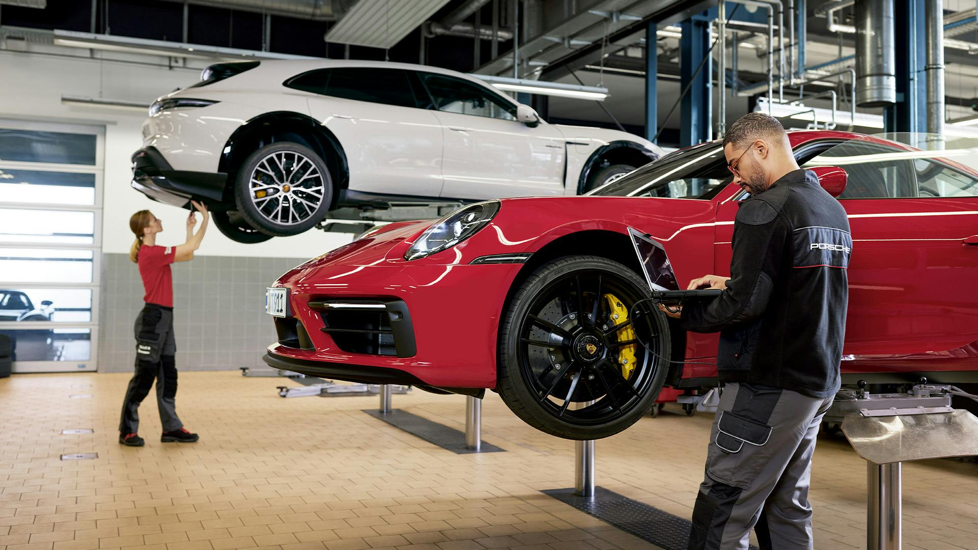 Two Porsche technicians working on cars in a modern workshop; a white Porsche SUV is elevated on a lift in the background, while a red Porsche sports car is being inspected in the foreground.
