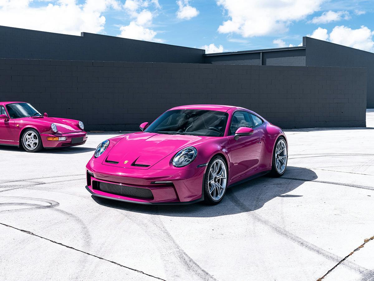 A 911 GT3 Touring and an older model both coloured in Rubystar Neo parking in different directions next to each other on a concrete flooring in front of a grey brick wall.