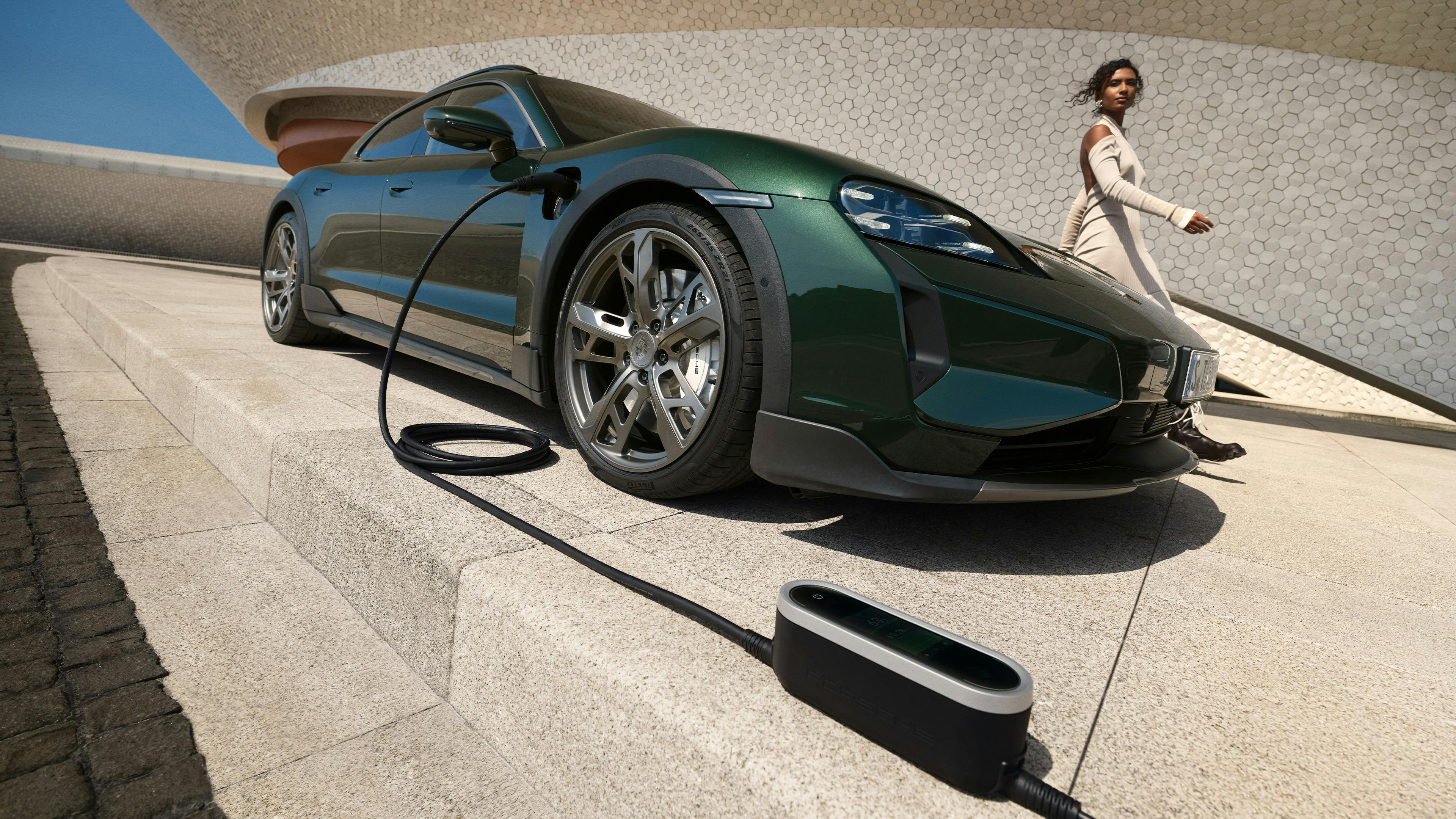 A Porsche Taycan Turbo Cross Turismo in front of a villa next to a Wallbox. A woman plugging in the plug.