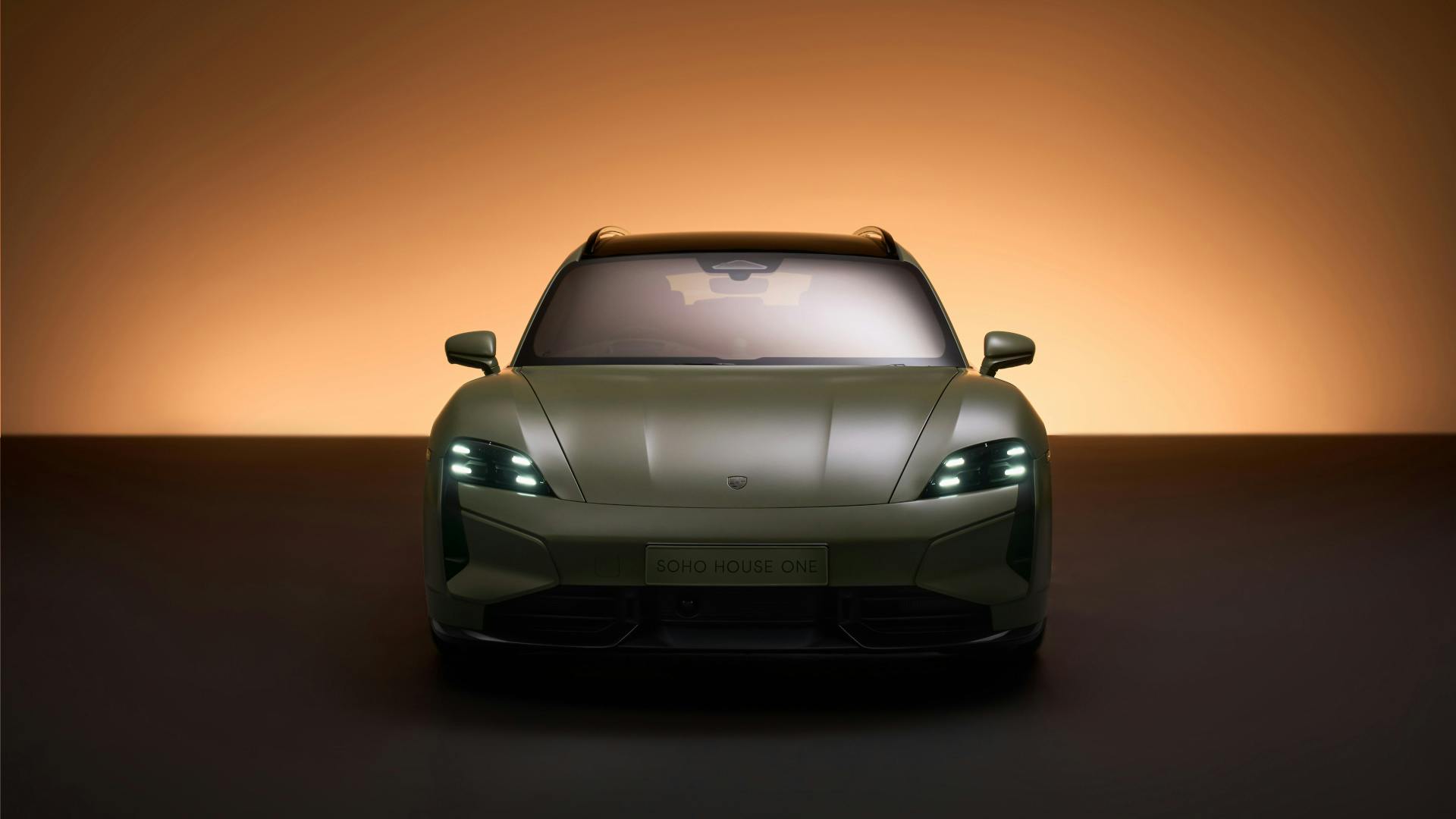 Front view of the Taycan Turbo S Soho House One in satin Greek Street Green, headlights lit, with Soho House One lettering on the front, against an amber studio backdrop.