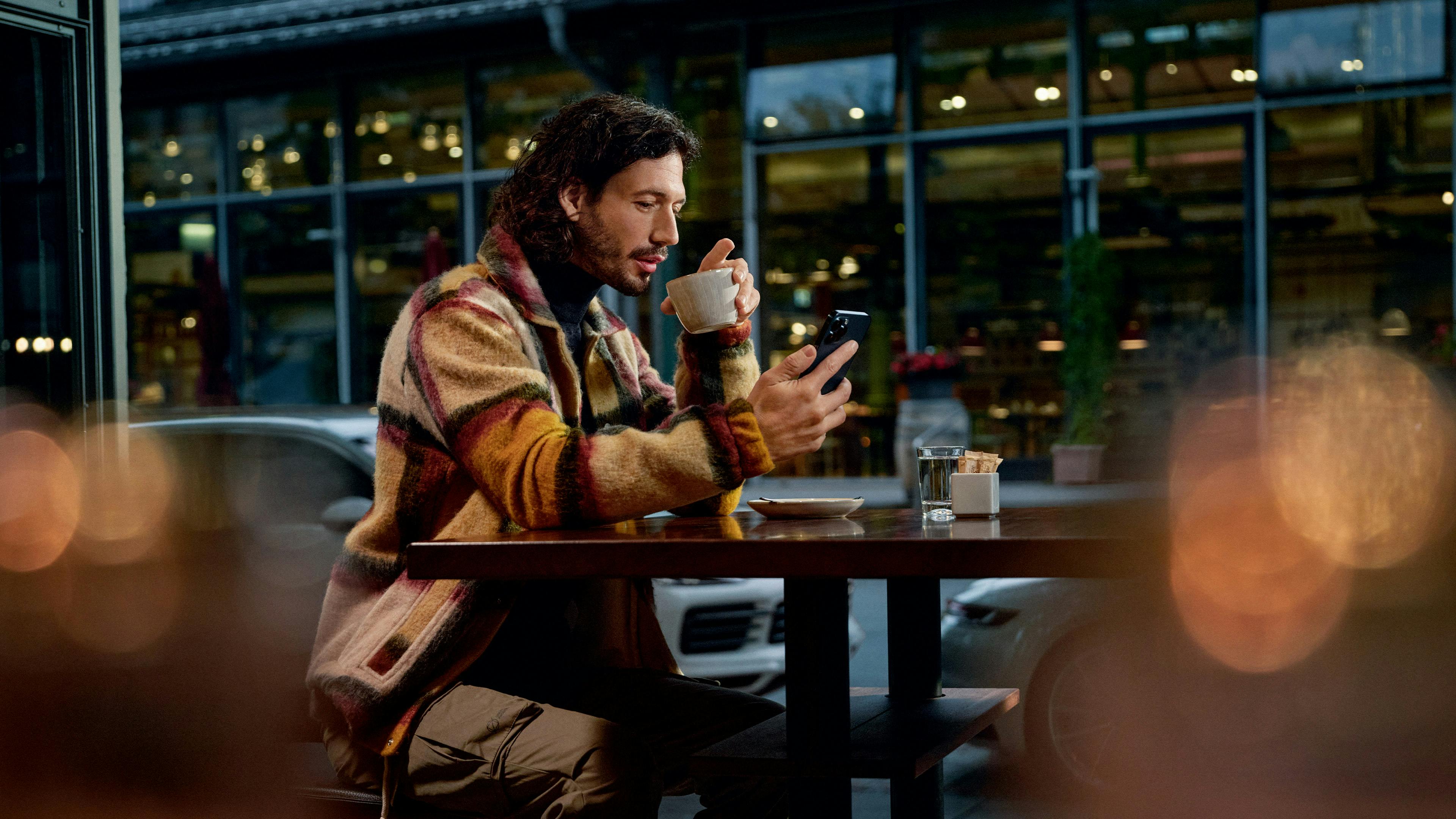 Un homme est assis dans un café. Il tient dans une main une tasse contenant une boisson chaude et dans l'autre un téléphone portable qu'il regarde.