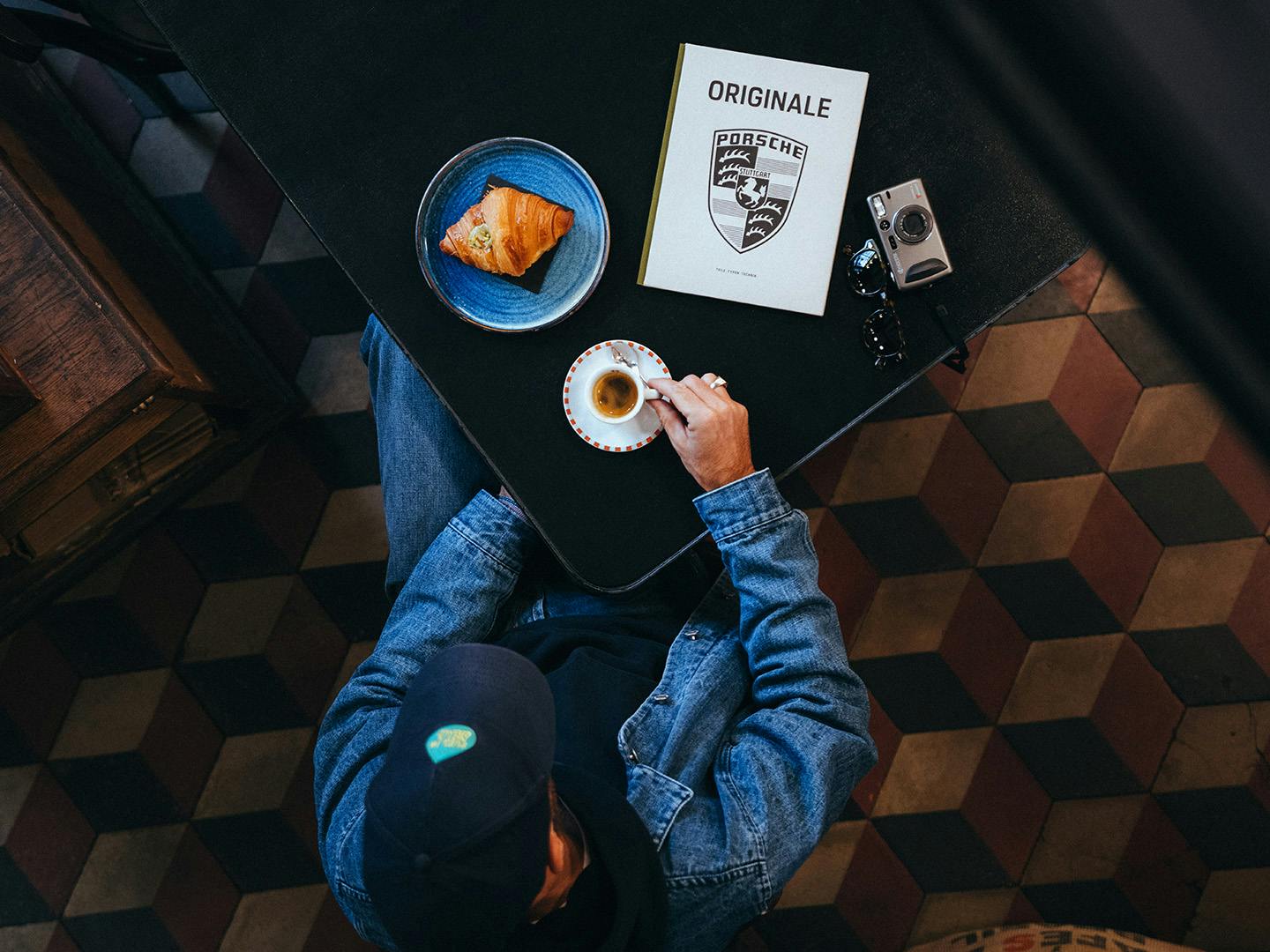 Andrea Vailetti sits at a table in a café with a copy of ORIGINALE 11. On the table are an espresso, a croissant, and a camera.