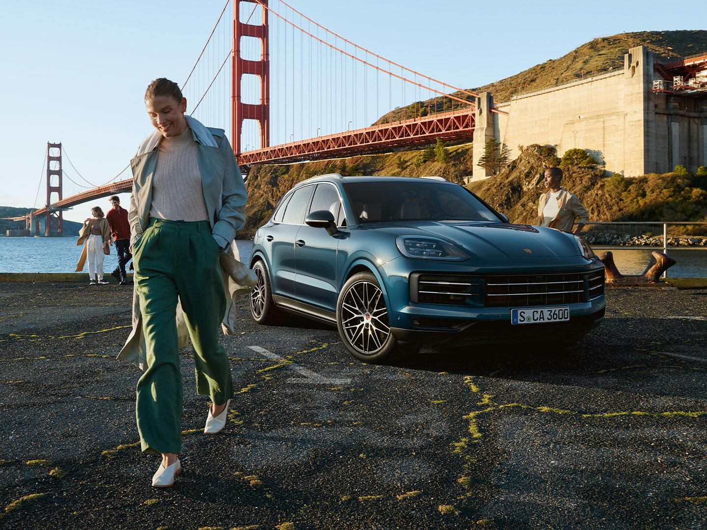 Una donna sorride mentre cammina vicino a una Porsche Cayenne blu, con il ponte Golden Gate sullo sfondo.
