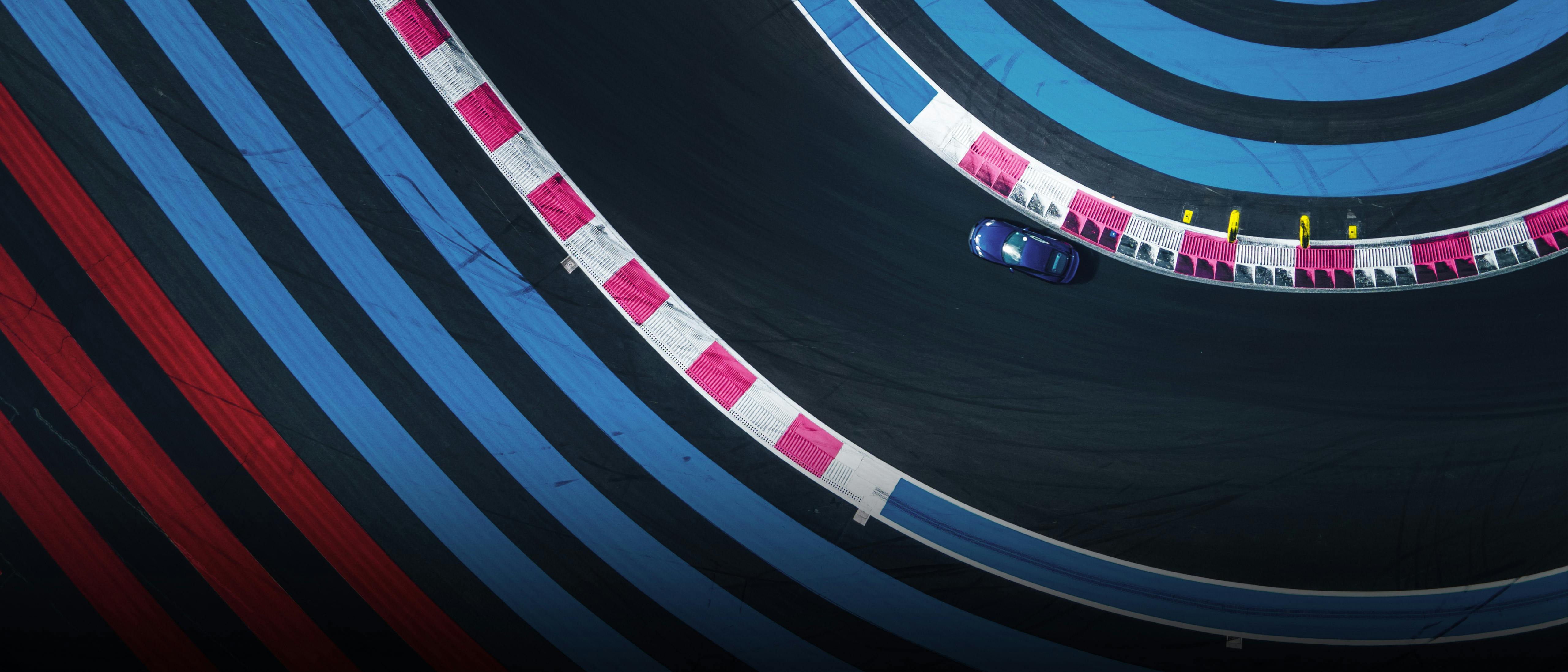 View from above of a Blue Porsche vehicle driving on a race track that is clearly bordered on the left and right by many blue and red guidelines.