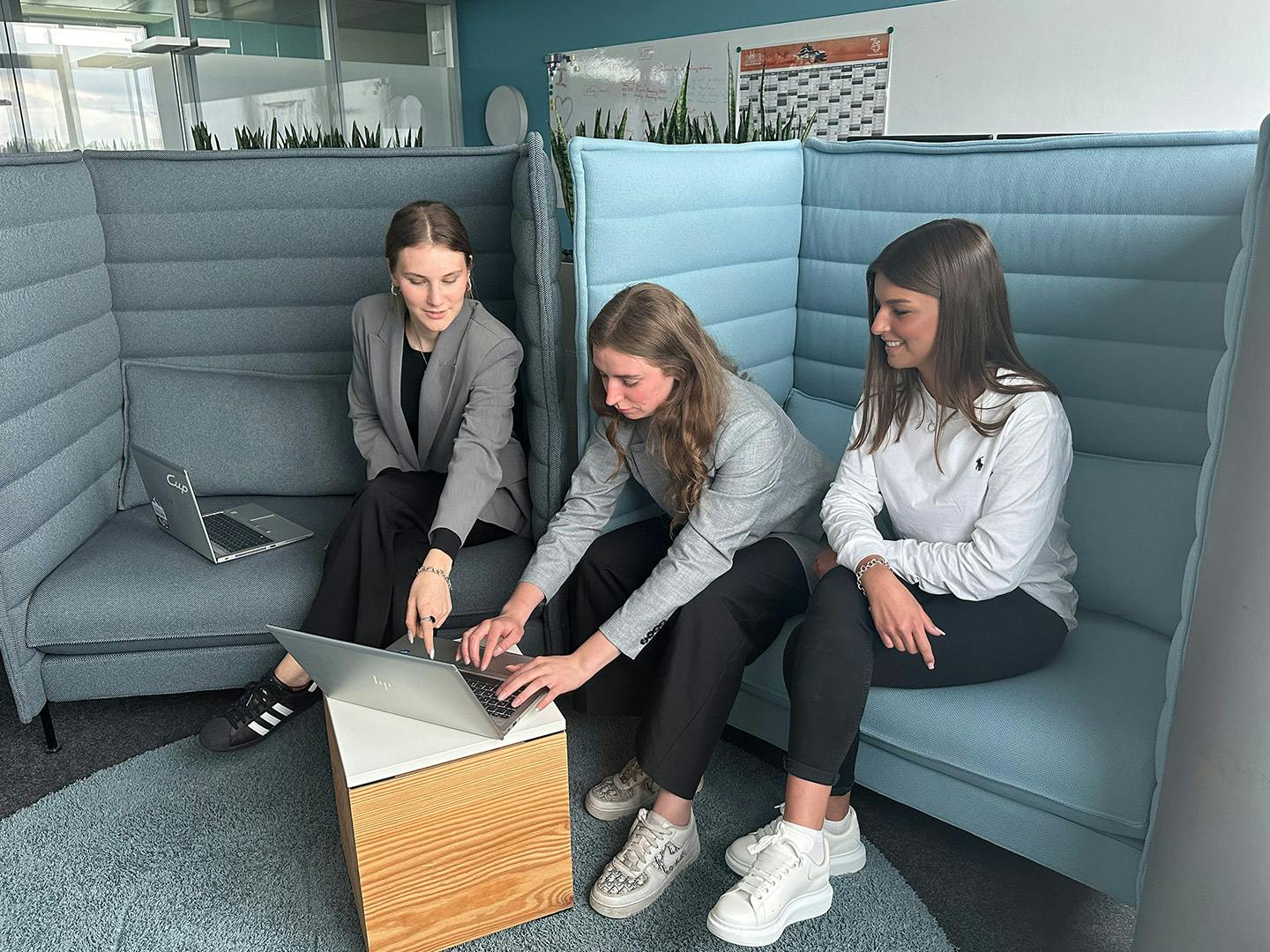 Drei Porsche Werkstudentinnen sitzen in grau-blauen Sesseln und arbeiten am Laptop.