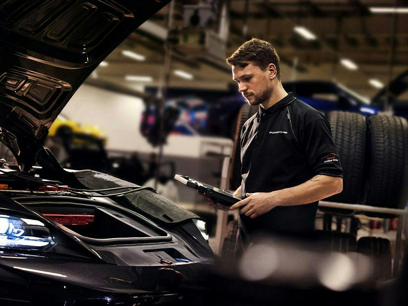 A male technician wearing a black Porsche-branded polo shirt stands in a professional garage. He is holding a diagnostic tablet and looking down at the front trunk area of a dark-coloured Porsche with its hood open. In the background, other cars and stacks of tires are visible.