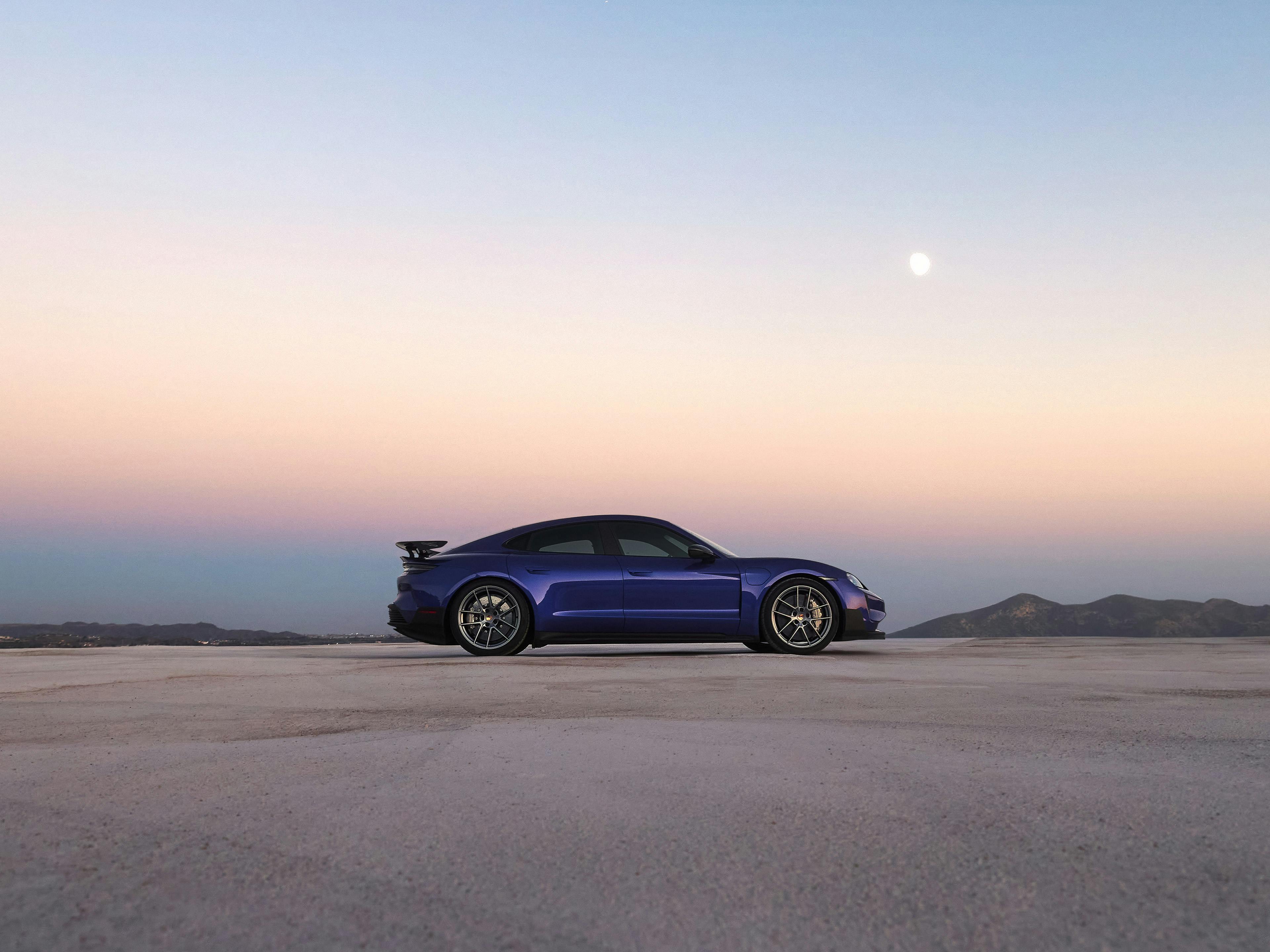 Ein blauer Sportwagen steht auf einer weiten, flachen Salzebene bei Sonnenuntergang. Im Hintergrund sind dunkle Hügel zu sehen, darüber ein klarer Himmel mit zarten Farbverläufen von Rosa über Violett zu Blau. Der Mond ist gut sichtbar am Himmel.