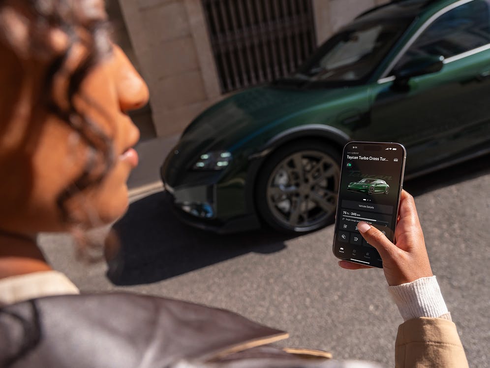 View over a woman's shoulder at a smartphone with the My Porsche app open, with the front of a Porsche Taycan Turbo Cross Turismo in the background.
