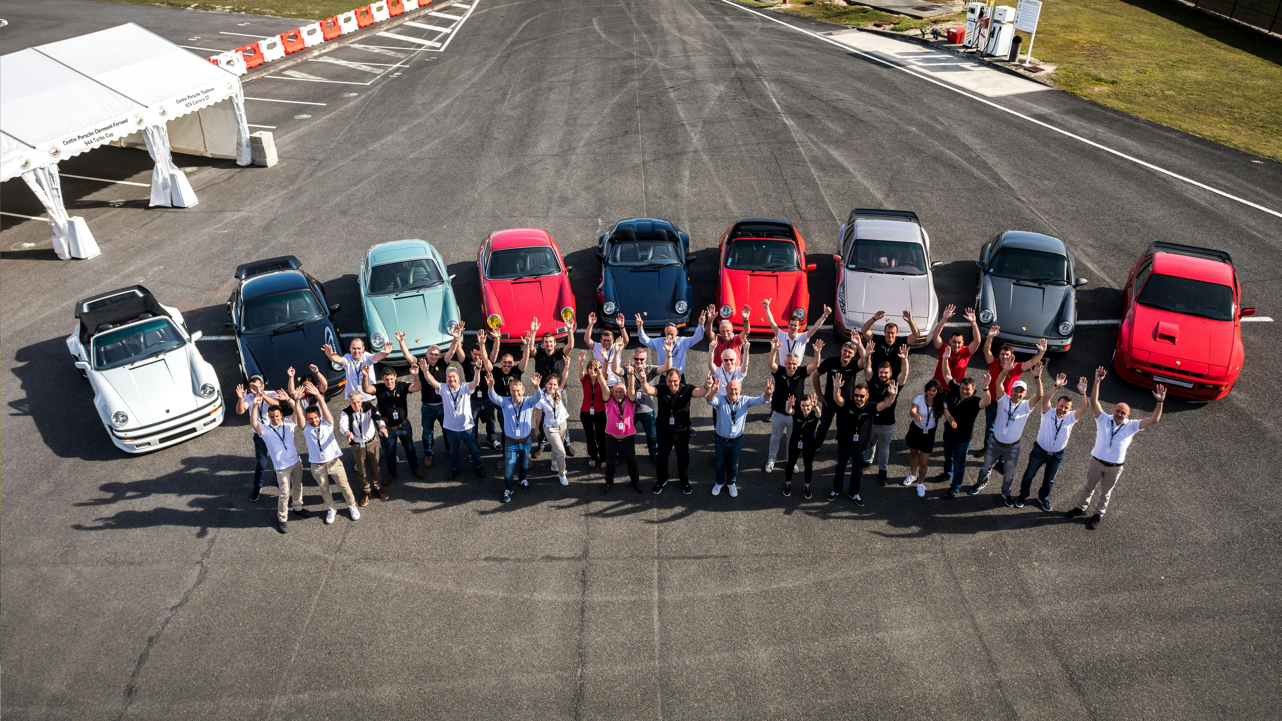 Les participants au Concours de Restauration se tiennent sur le circuit. Derrière eux, les neuf voitures participantes sont alignées les unes à côté des autres.