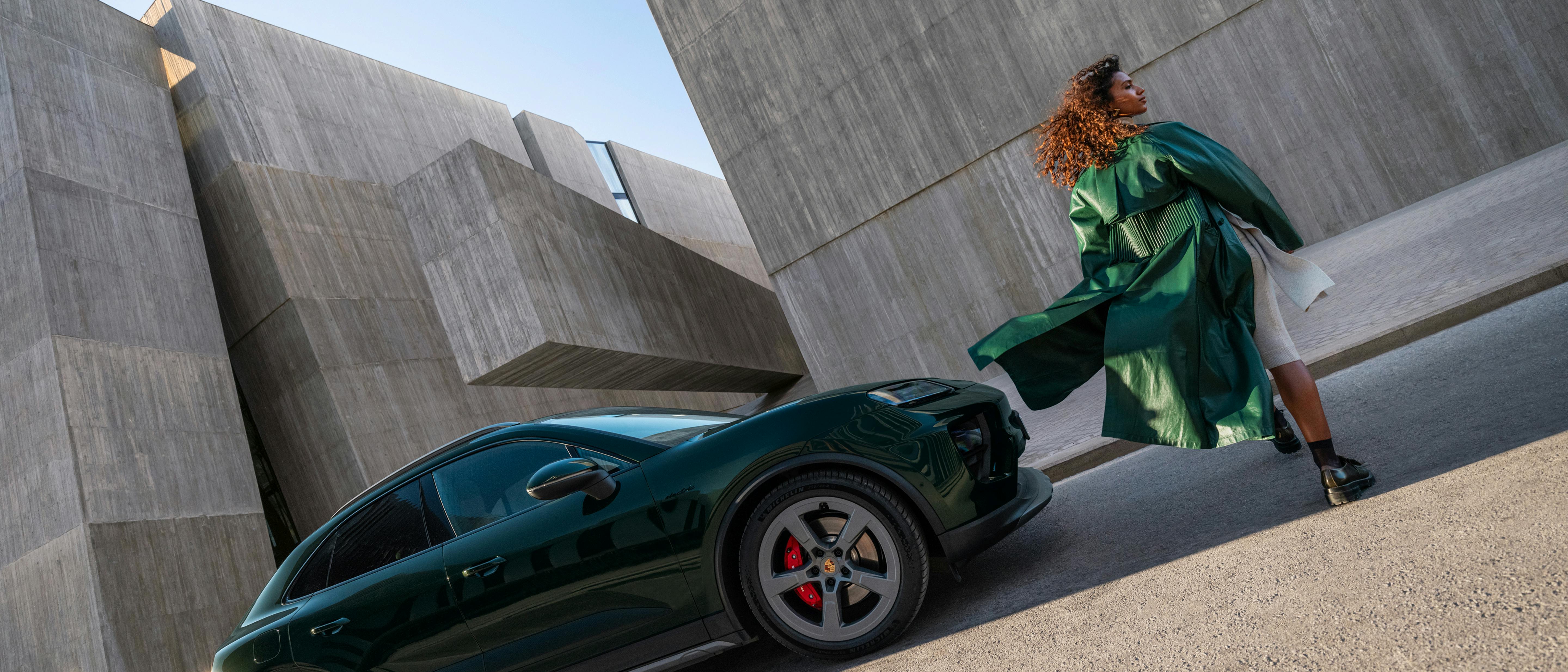 A Porsche Macan 4S in Oak Green Metallic stands in front of concrete walls. A woman in a green coat walks by.