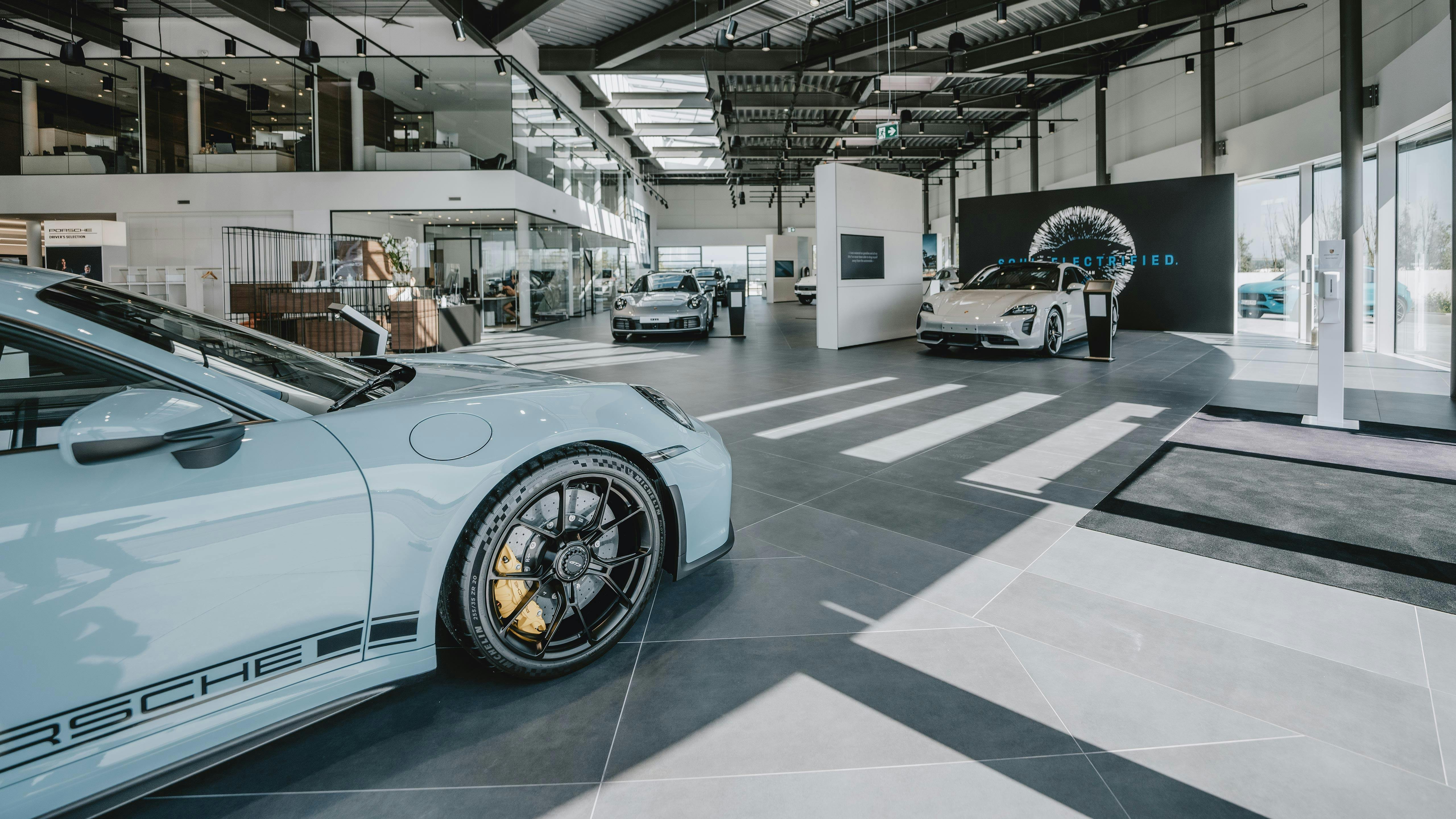 Intérieur du Porsche Centre Roost mettant en avant différents modèles Porsche. Grand espace ouvert avec un sol en carrelage gris et une lumière naturelle traversant les fenêtres.