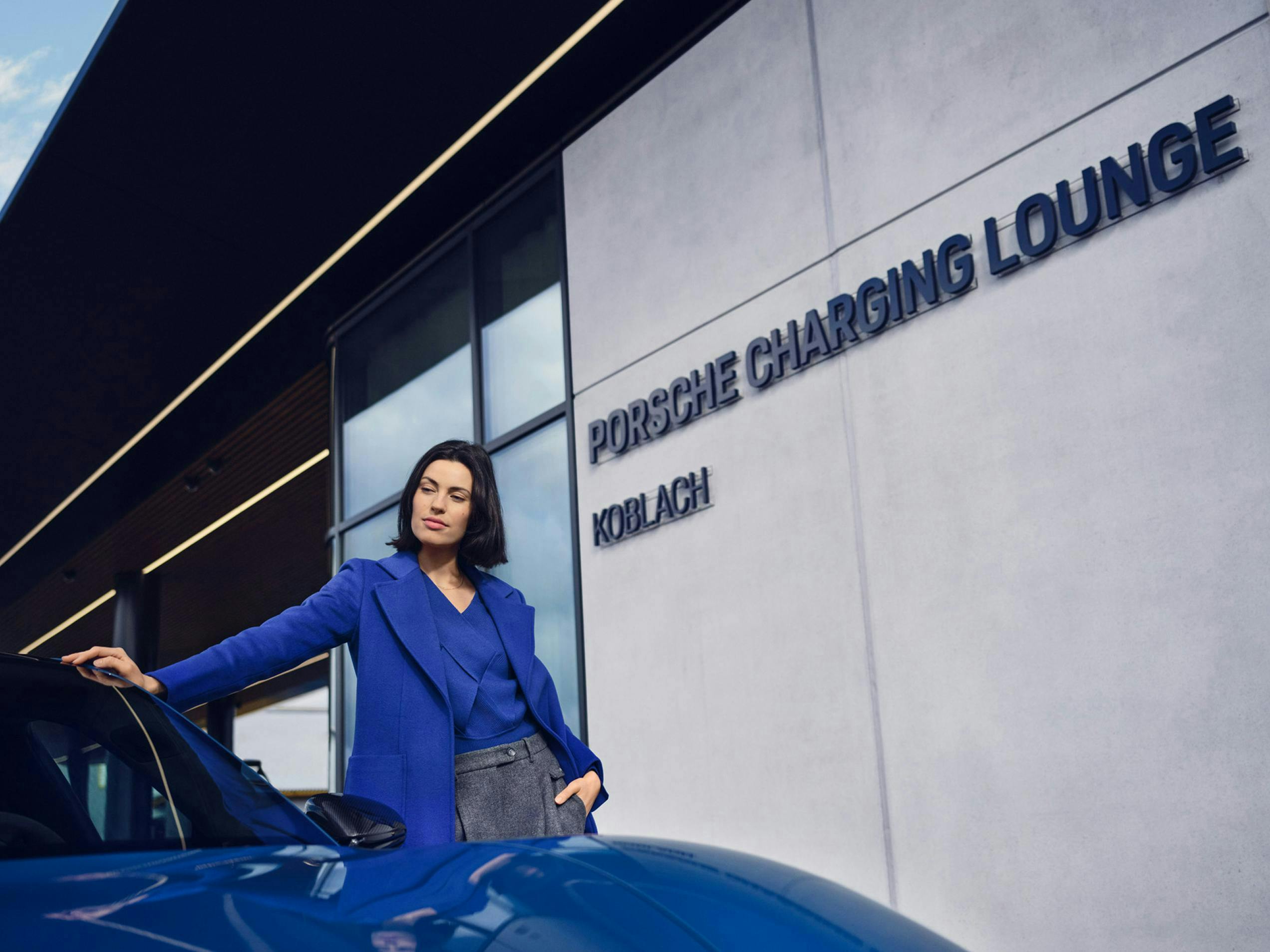 Kobieta w niebieskiej marynarce oparta o niebieskie Porsche przed drzwiami Porsche Charging Lounge w Koblach.